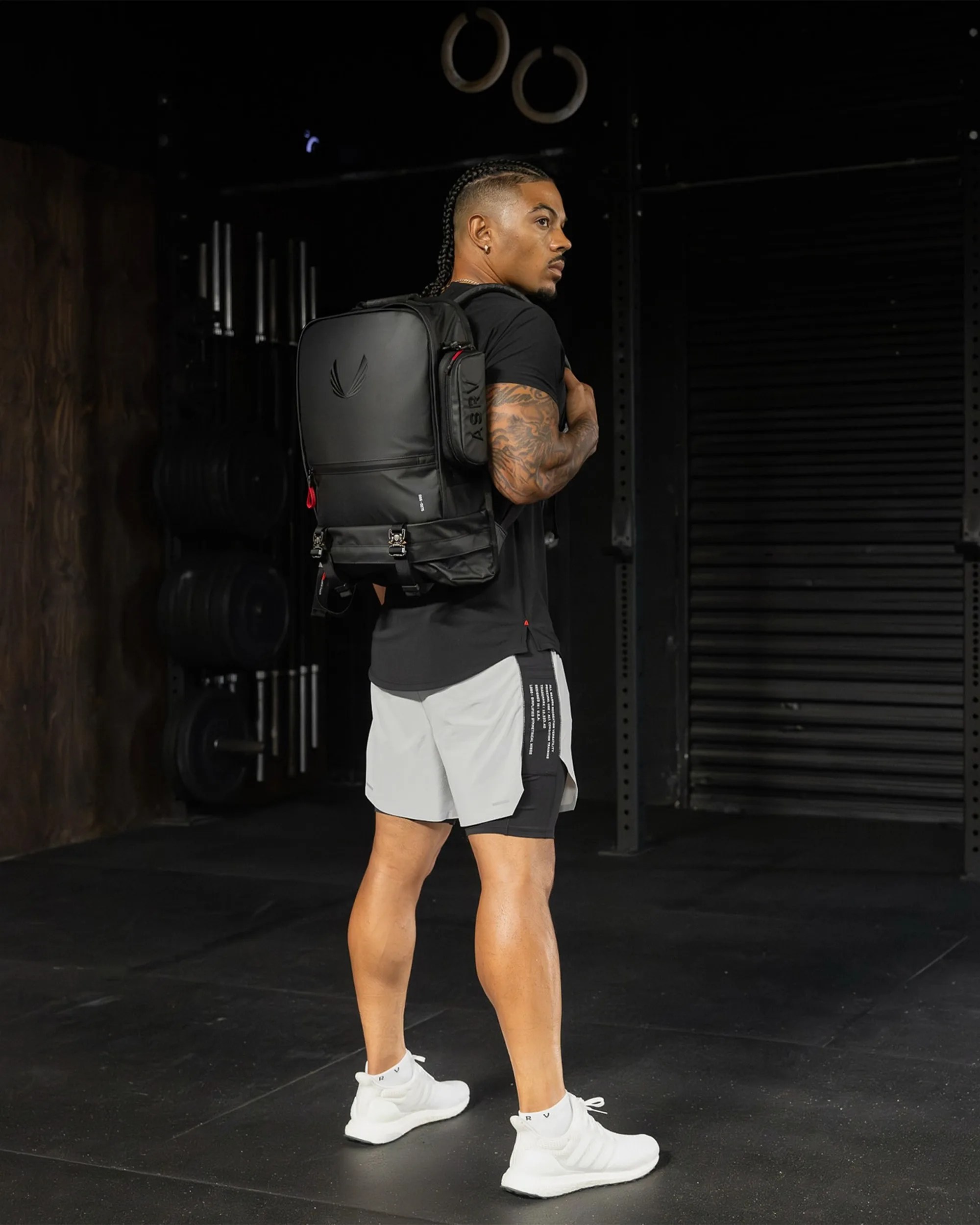 Athletic person wearing white sneakers, black and gray shorts, black shirt, and carrying a black backpack in a gym.