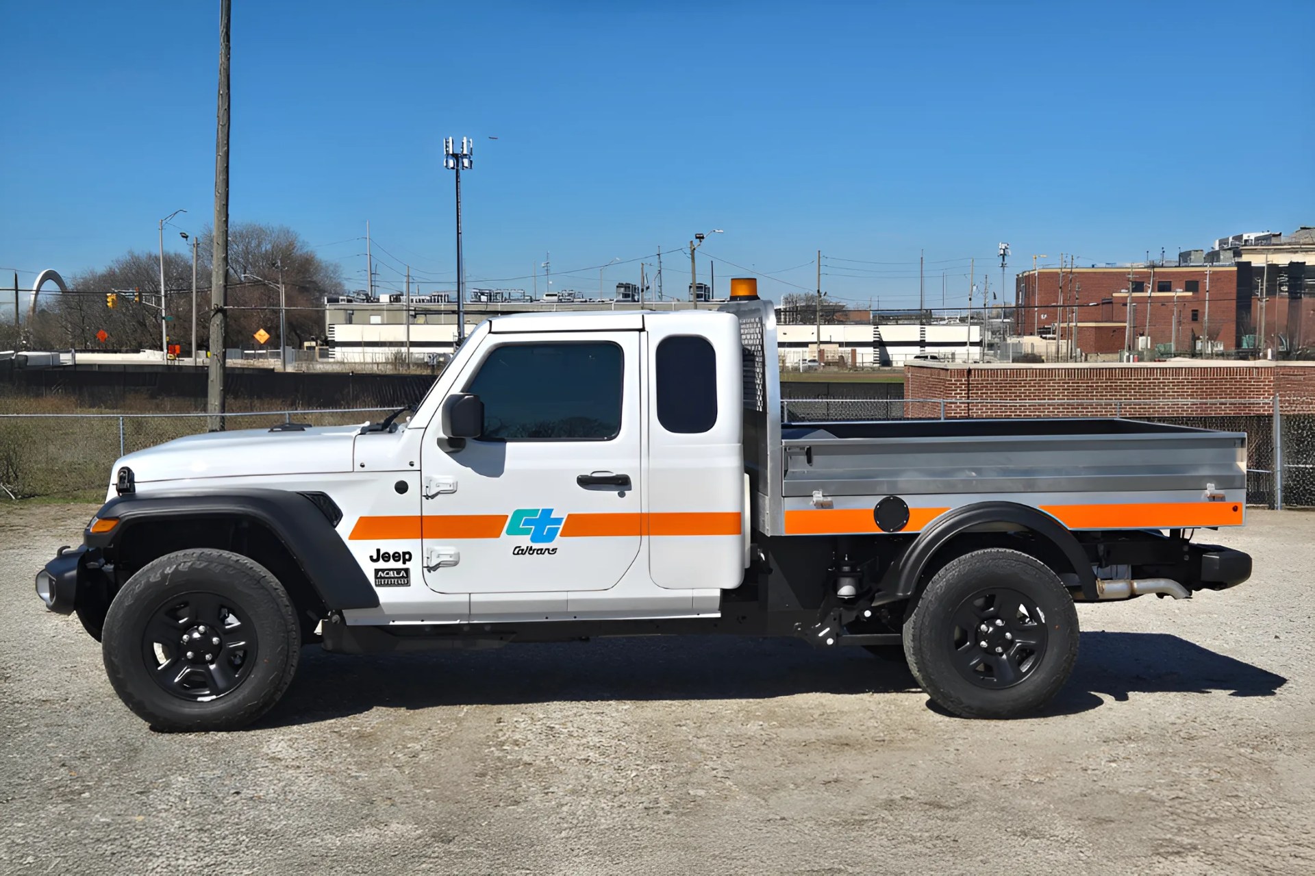 White Jeep pickup truck with orange stripe and Caltrans logo parked on gravel lot.