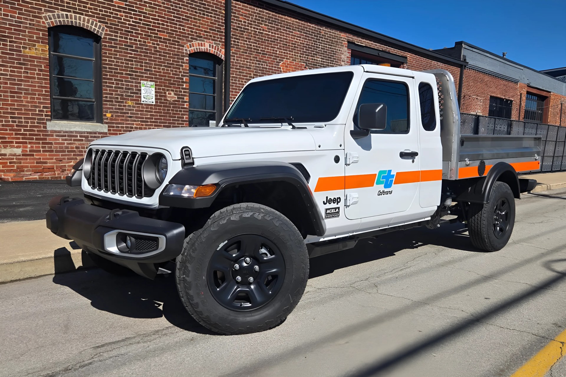White Jeep Gladiator pickup truck with Caltrans logo and orange stripe parked on a street in front of a brick building.