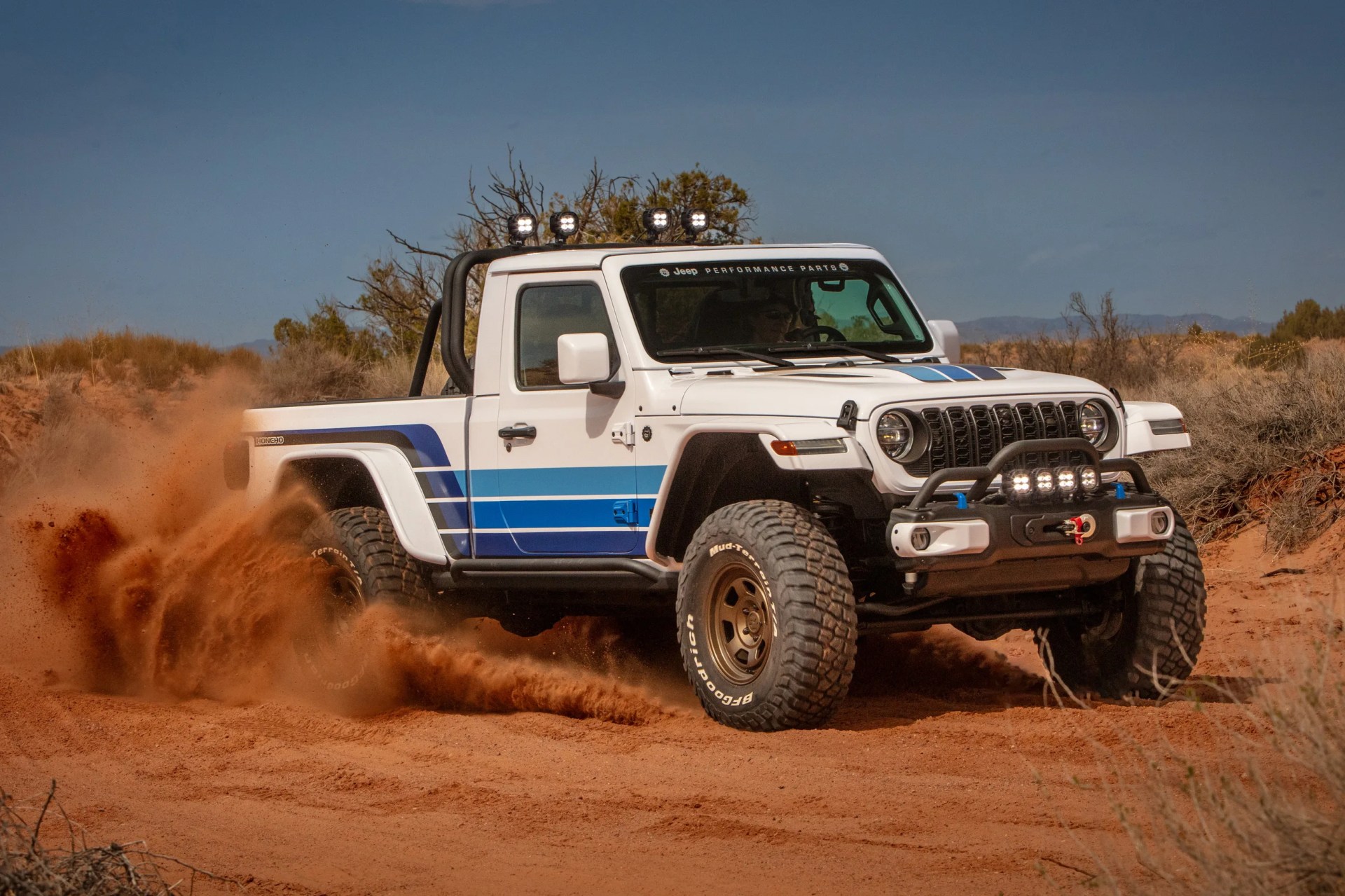 White Jeep Gladiator pickup with blue stripes kicking up red dirt in a desert landscape.
