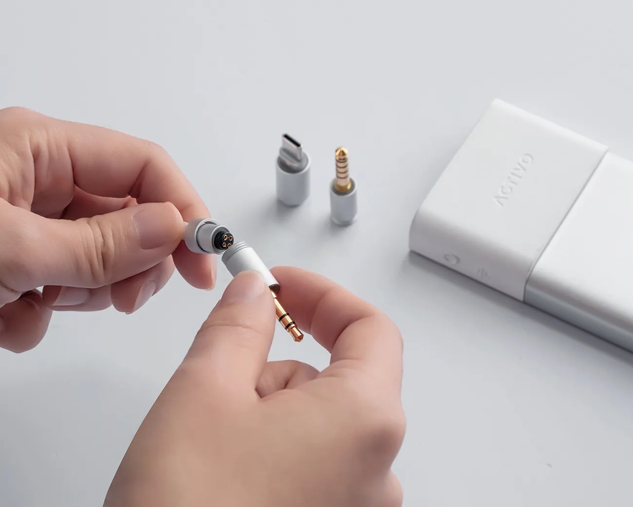 Hands holding a white magnetic adapter with gold 3.5mm audio jack pins near a white device and two other adapters on a gray surface.