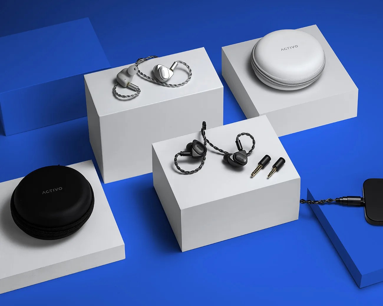 Two pairs of in-ear wired earphones, one silver and one black, with braided cables, two round carrying cases labeled "ACTIVO," and two audio adapters on white and blue blocks.
