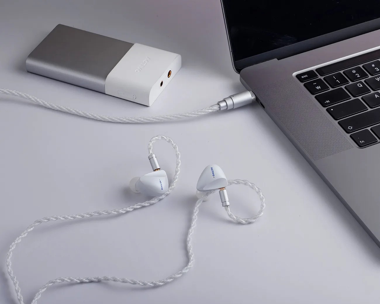 White braided wired in-ear monitors labeled "ACTIVO" connected to a silver and white portable DAC/amp and a gray laptop.