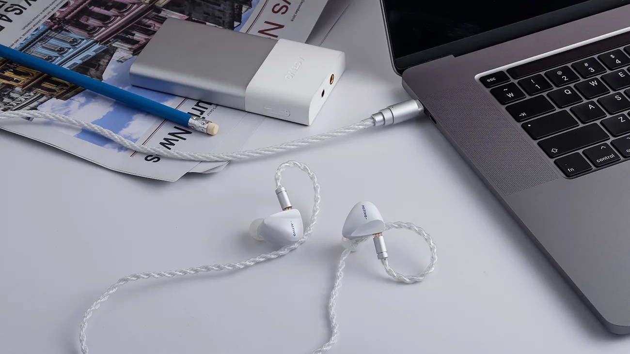 White wired in-ear monitors with braided cables connected to a silver laptop on a white desk.