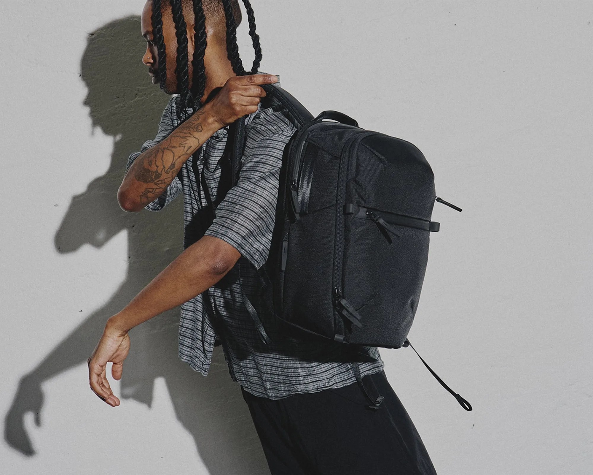 Black rectangular backpack with multiple zippered compartments worn over one shoulder by a person in a striped shirt.