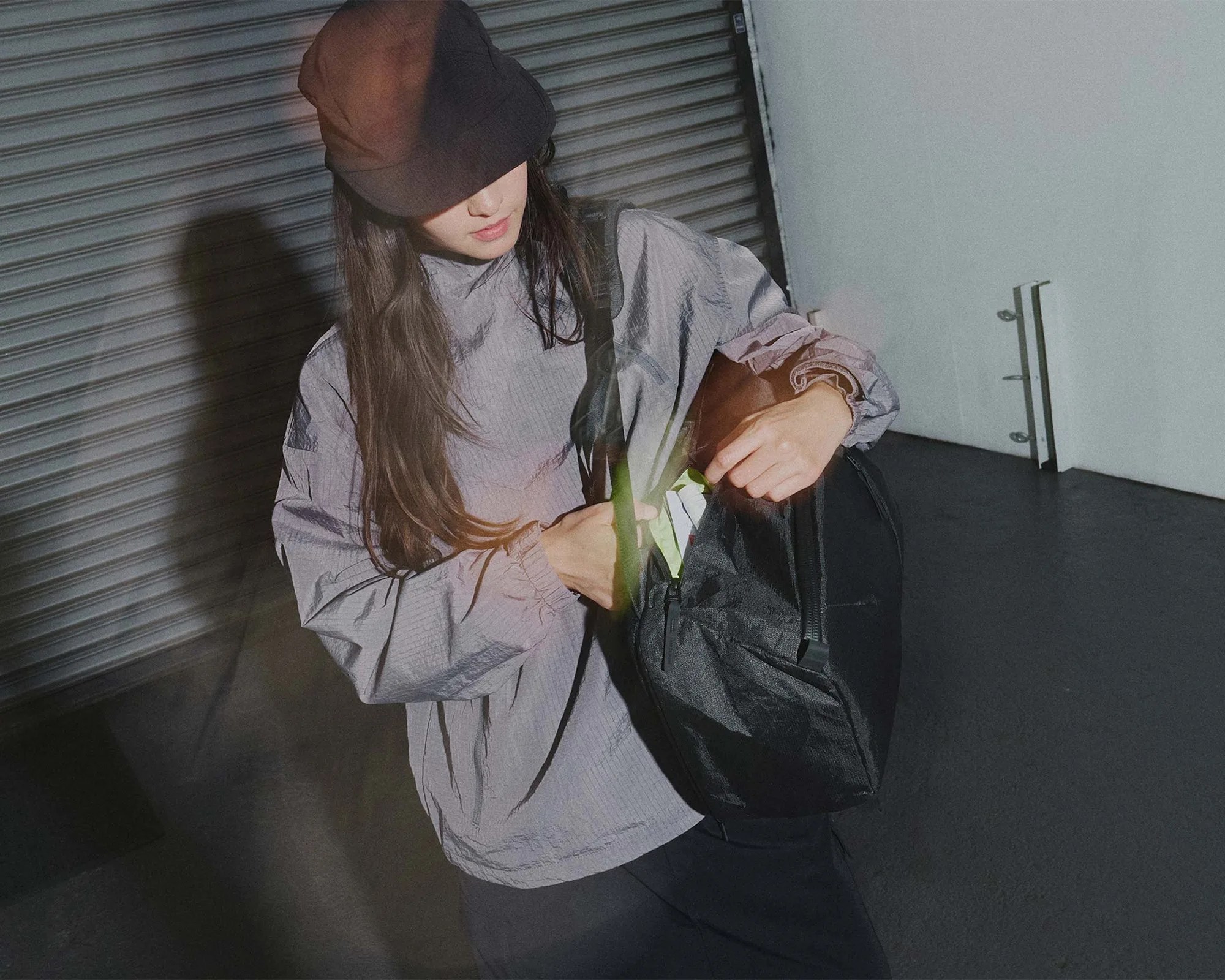 Person wearing a gray long-sleeve shirt and black cap, placing a green item into a black sling bag.