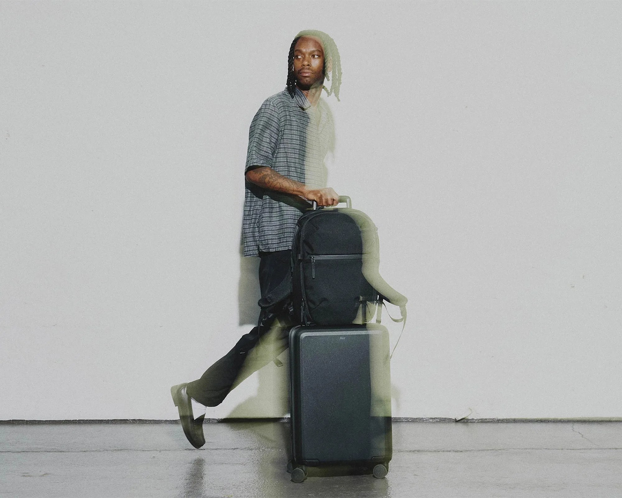 Person walking with a black backpack on top of a black rolling suitcase against a plain light wall.