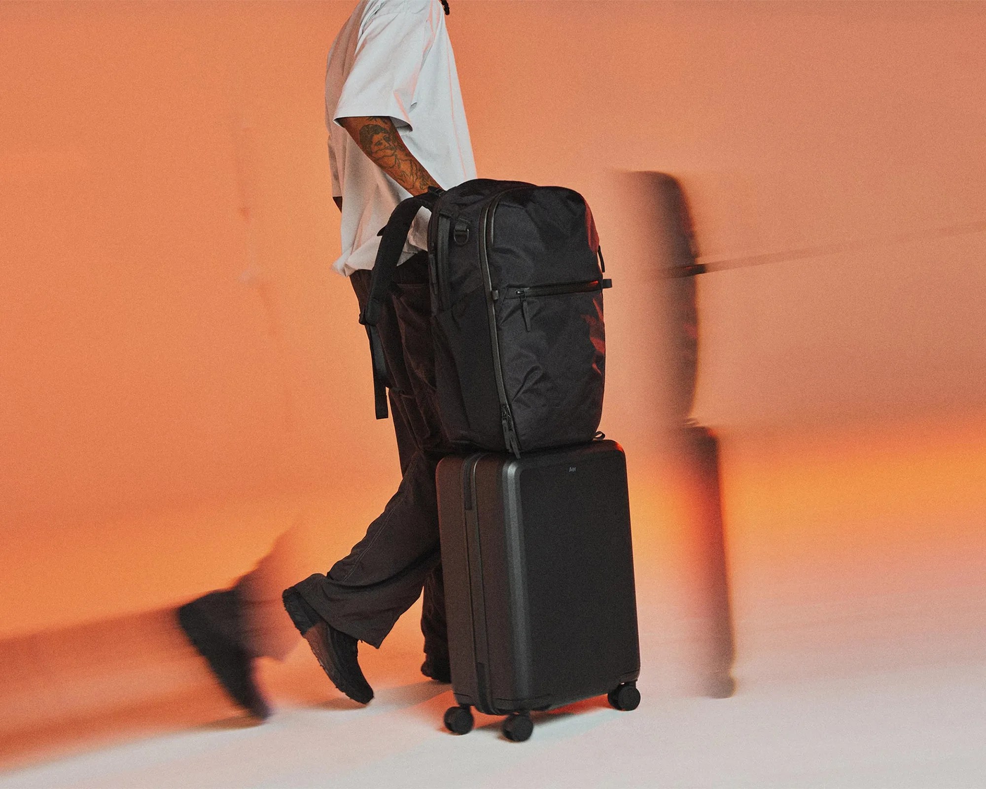 Person pulling a black hard-shell suitcase with a black backpack on top against an orange background.