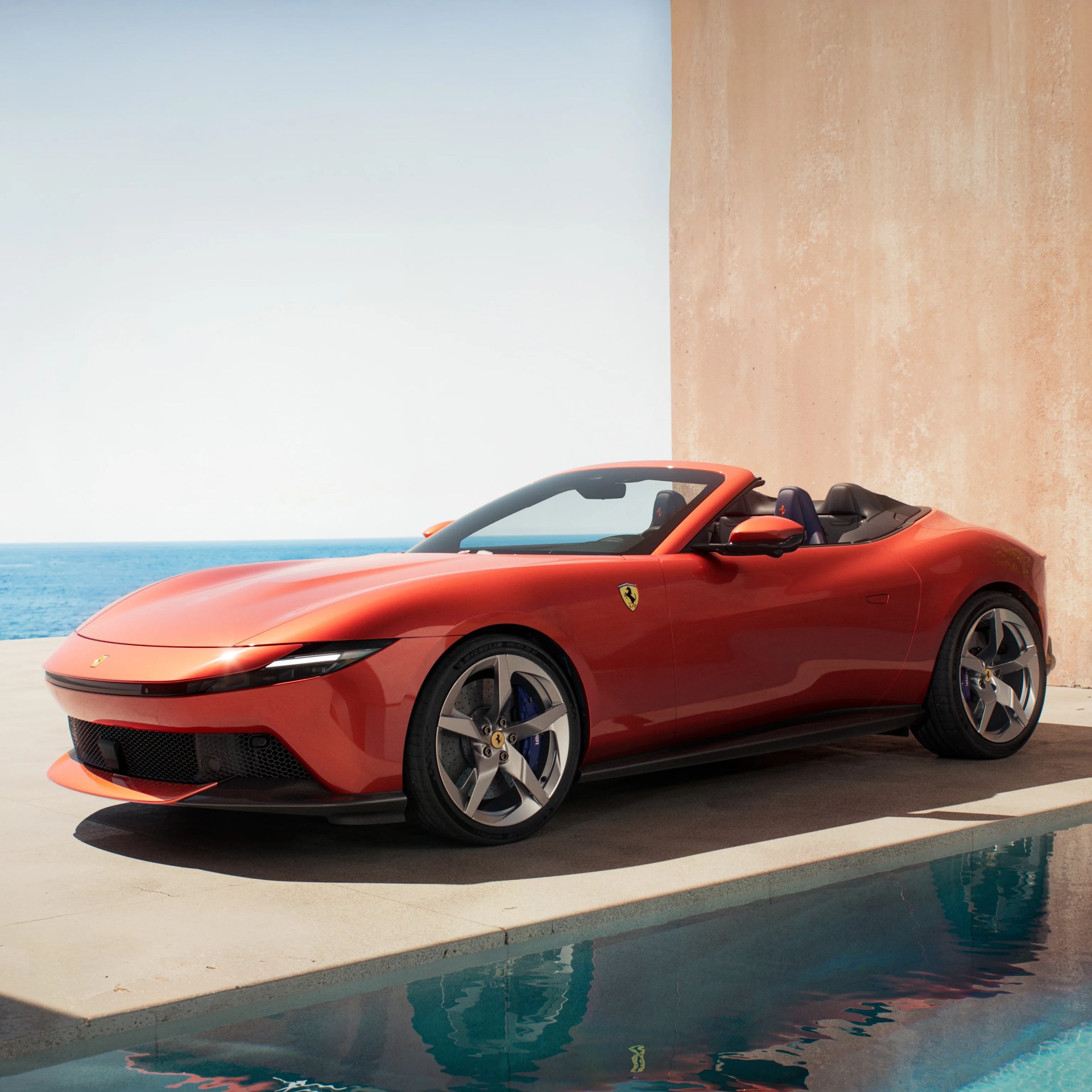 Red Ferrari convertible sports car parked near a pool with ocean in the background.