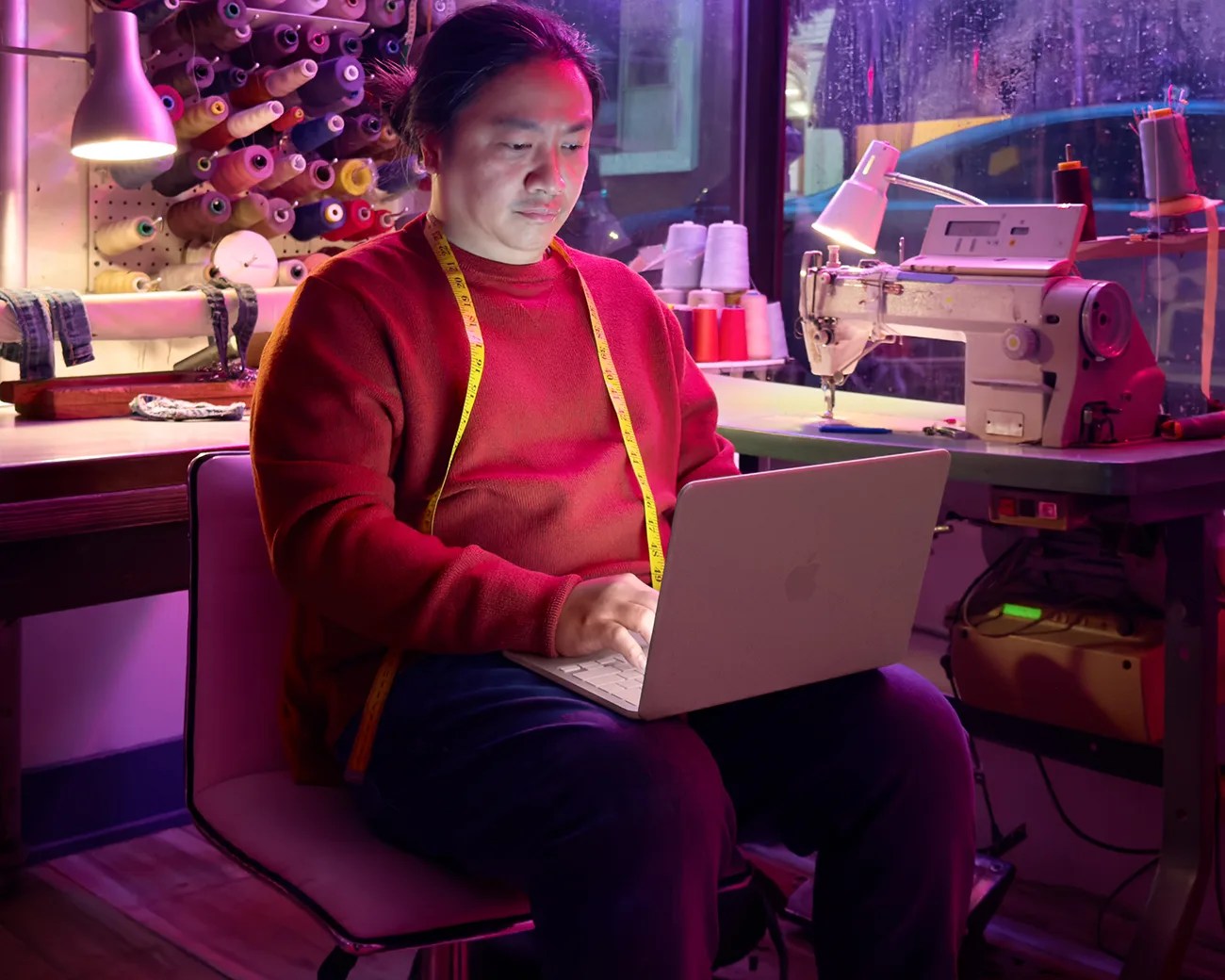 Person in a red sweater with a yellow measuring tape around their neck using a silver laptop in a sewing workshop.
