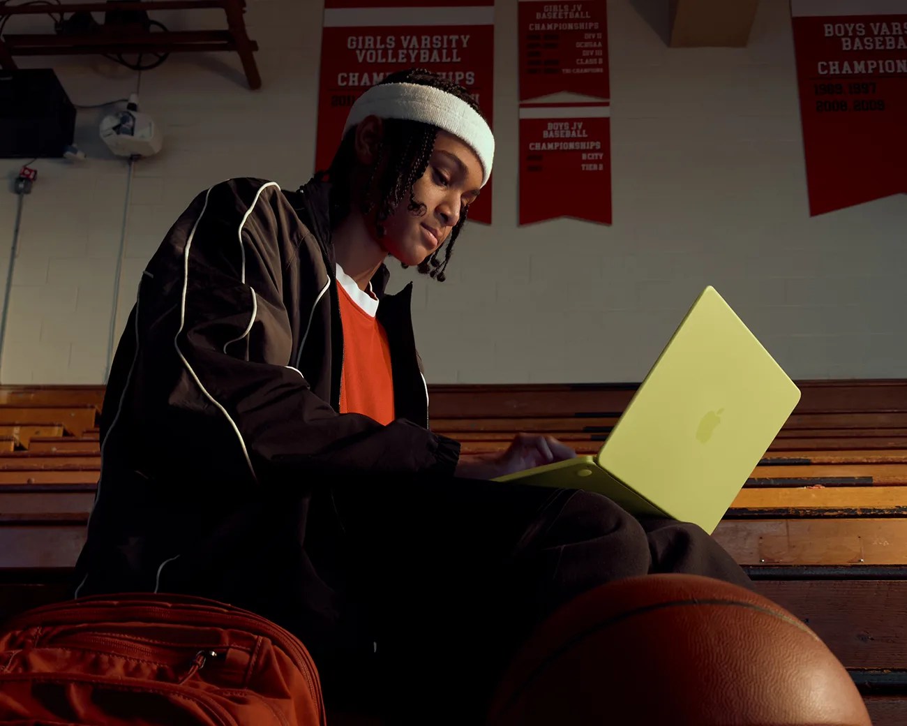 Person wearing a white headband and black jacket using a yellow Apple laptop in a gym with basketball and backpack nearby.
