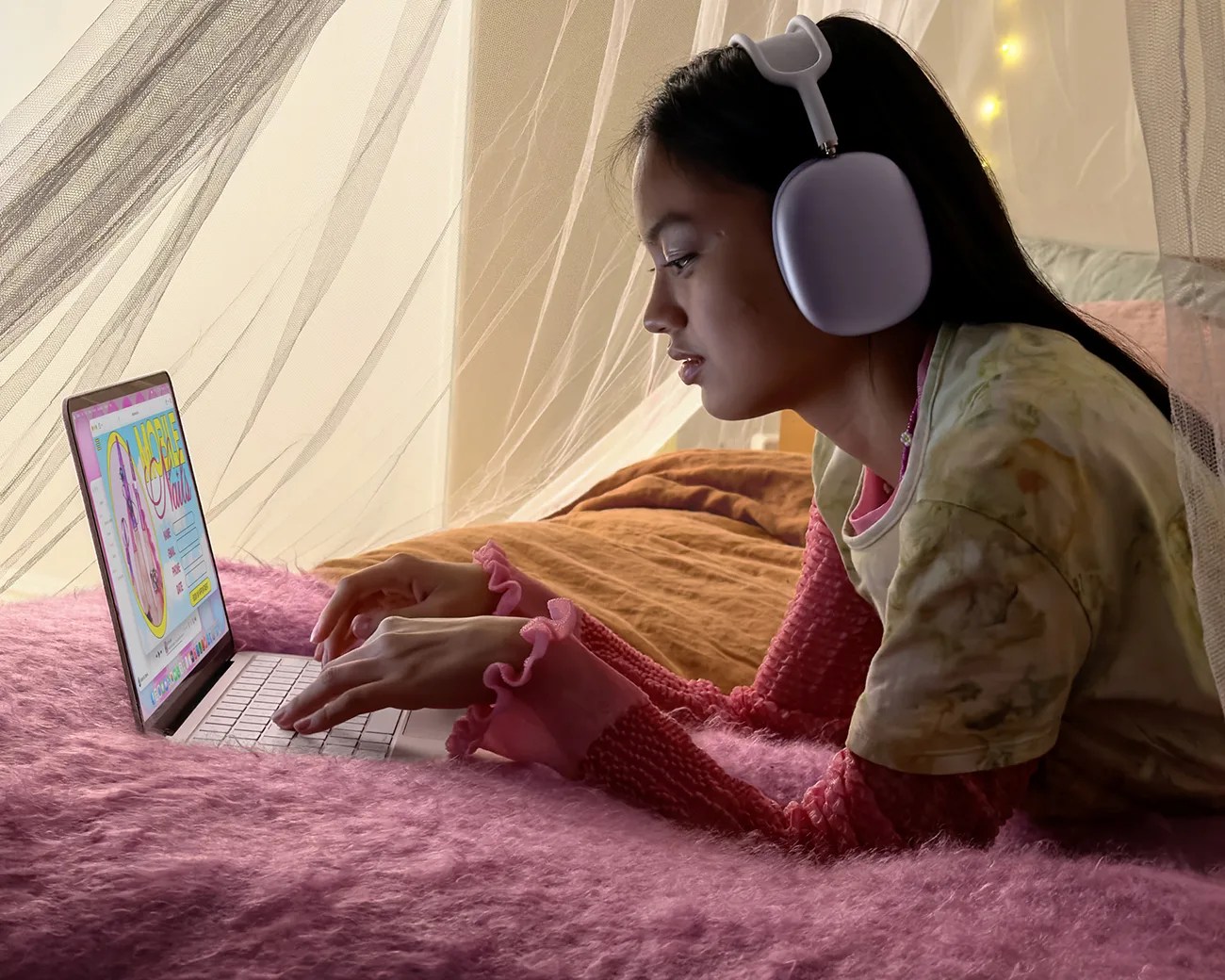 Person wearing white over-ear headphones and a layered pink and yellow top, using a laptop on a pink fuzzy blanket.