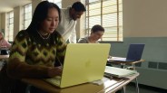 Student typing on a yellow Apple laptop at a classroom desk with notebooks and a pen nearby.
