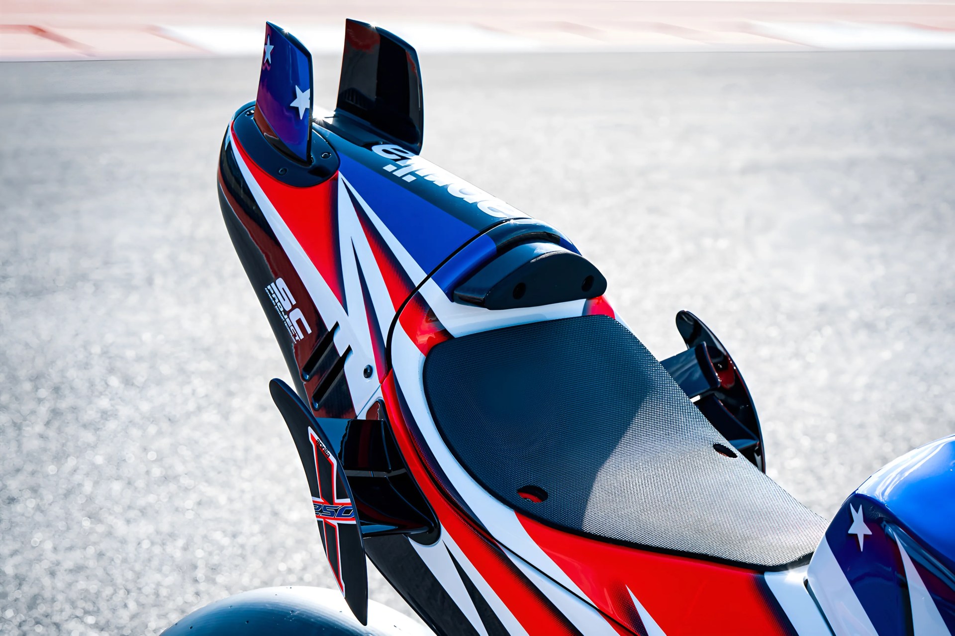 Rear section of a red, white, and blue racing motorcycle with aerodynamic winglets and a textured black seat.