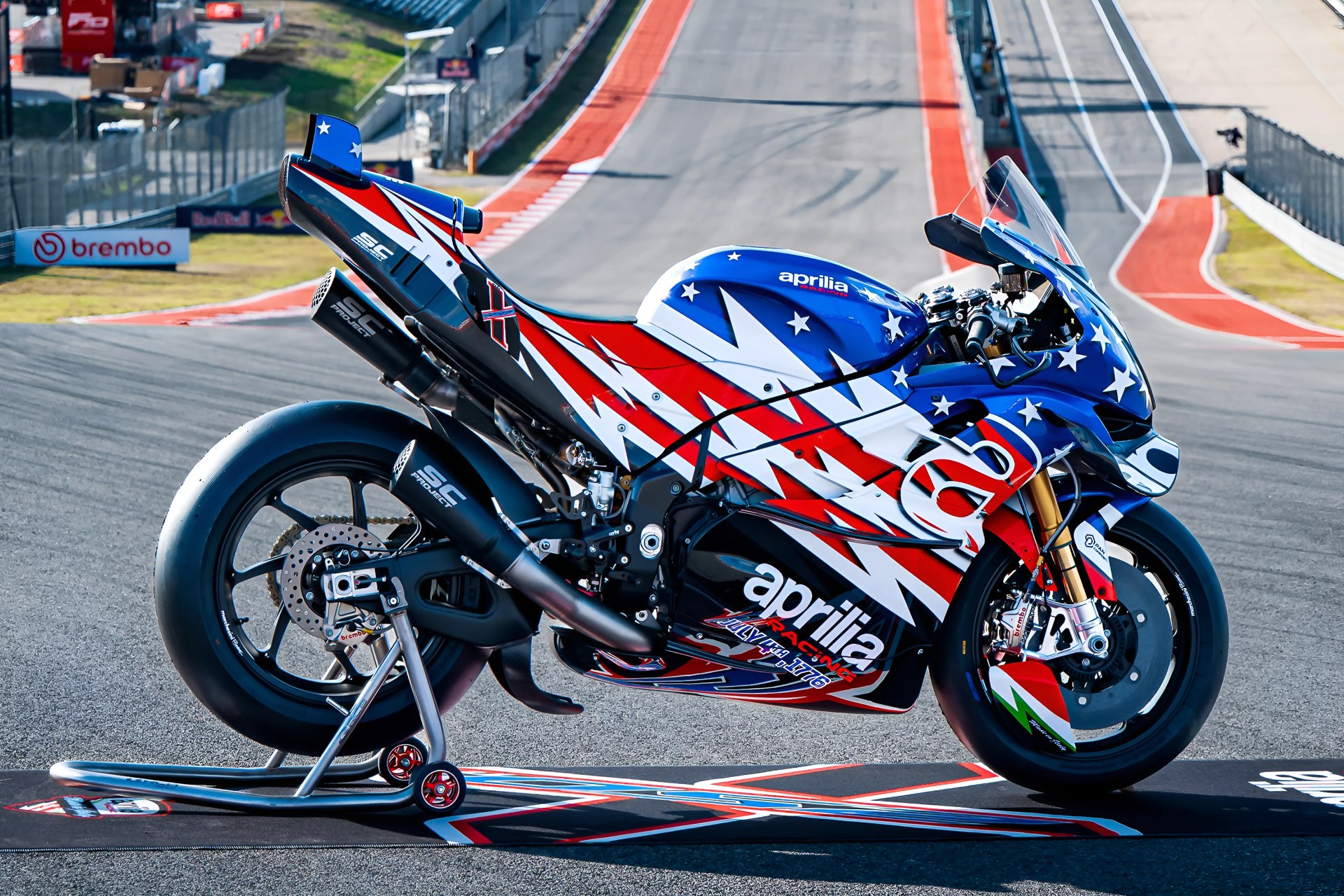 Blue, red, and white Aprilia racing motorcycle with stars and stripes design on a racetrack.