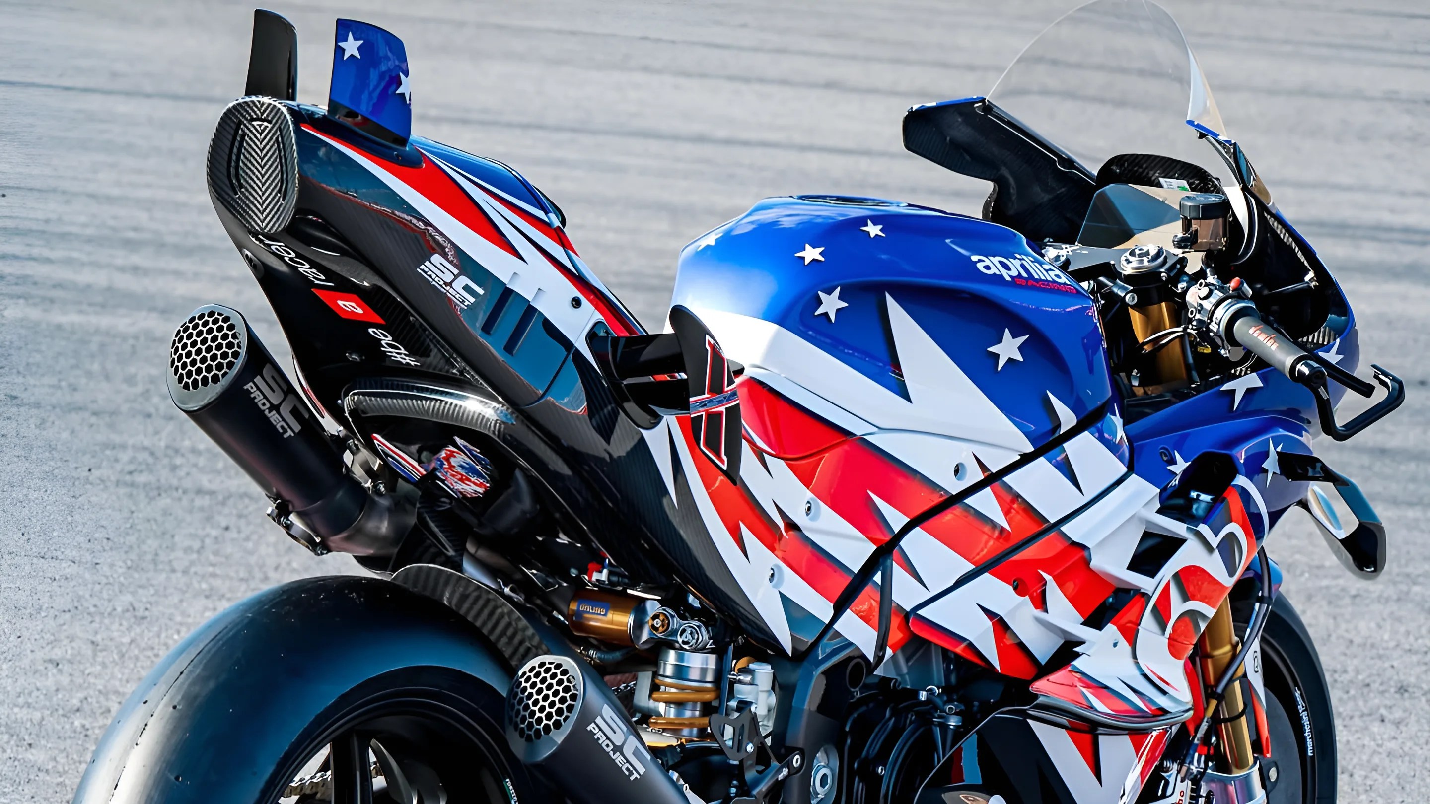 Blue, red, and white Aprilia racing motorcycle with SC Project dual exhausts on a racetrack.