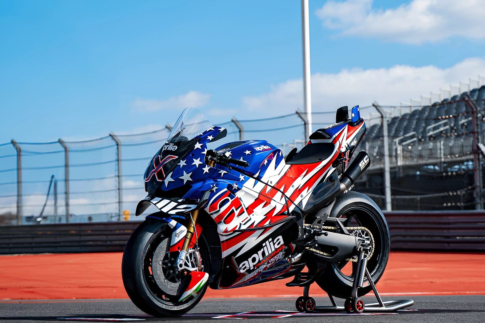 Blue, red, and white Aprilia racing motorcycle with stars and stripes design on a racetrack.