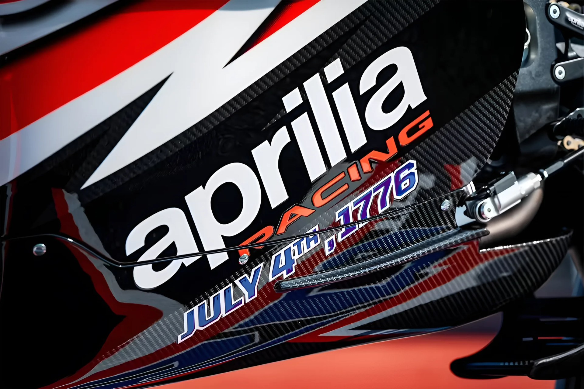 Close-up of a black carbon fiber motorcycle fairing with "aprilia RACING" and "JULY 4TH, 1776" decals.