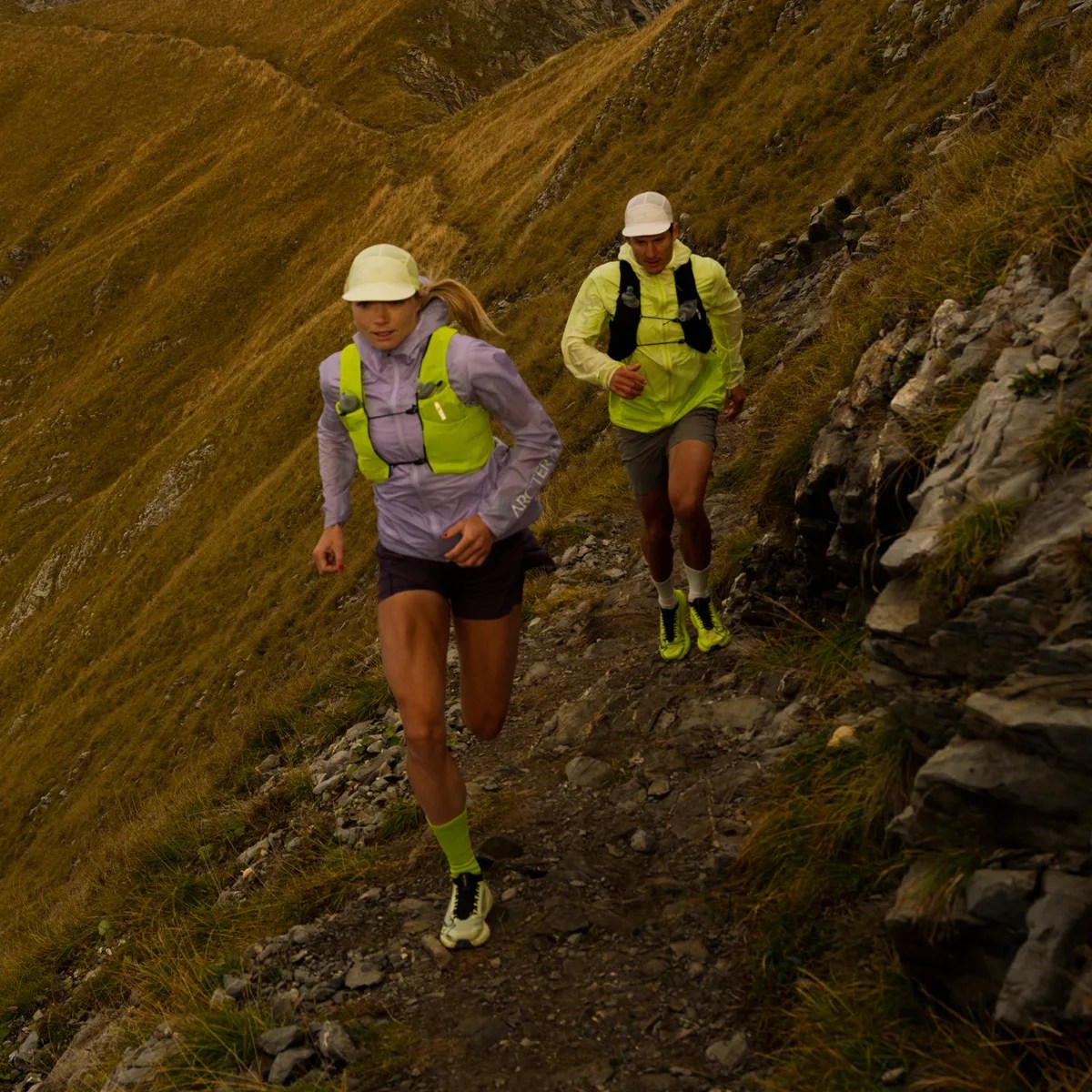 Two trail runners wearing neon yellow vests and caps running uphill on a rocky mountain path.