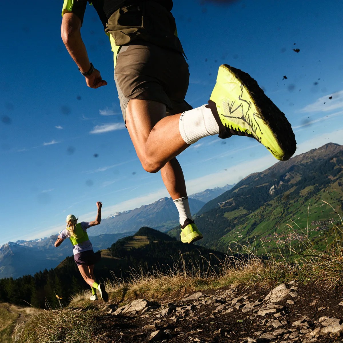 Two trail runners wearing neon yellow shoes and gear running on a rocky mountain path with a scenic valley and mountains in the background.