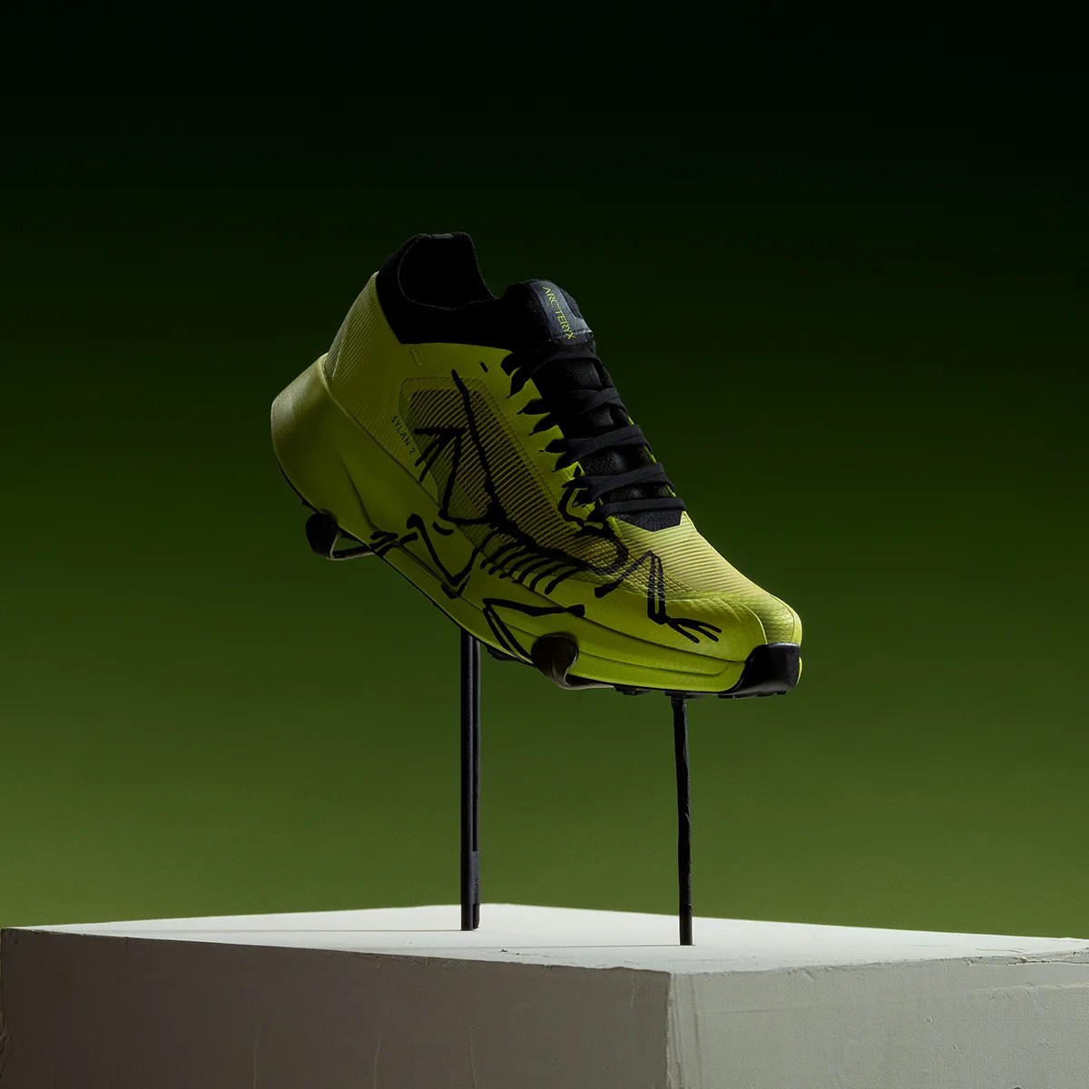 Yellow-green cleat shoe with black skeletal graphic and black laces displayed on black stands against a green background.