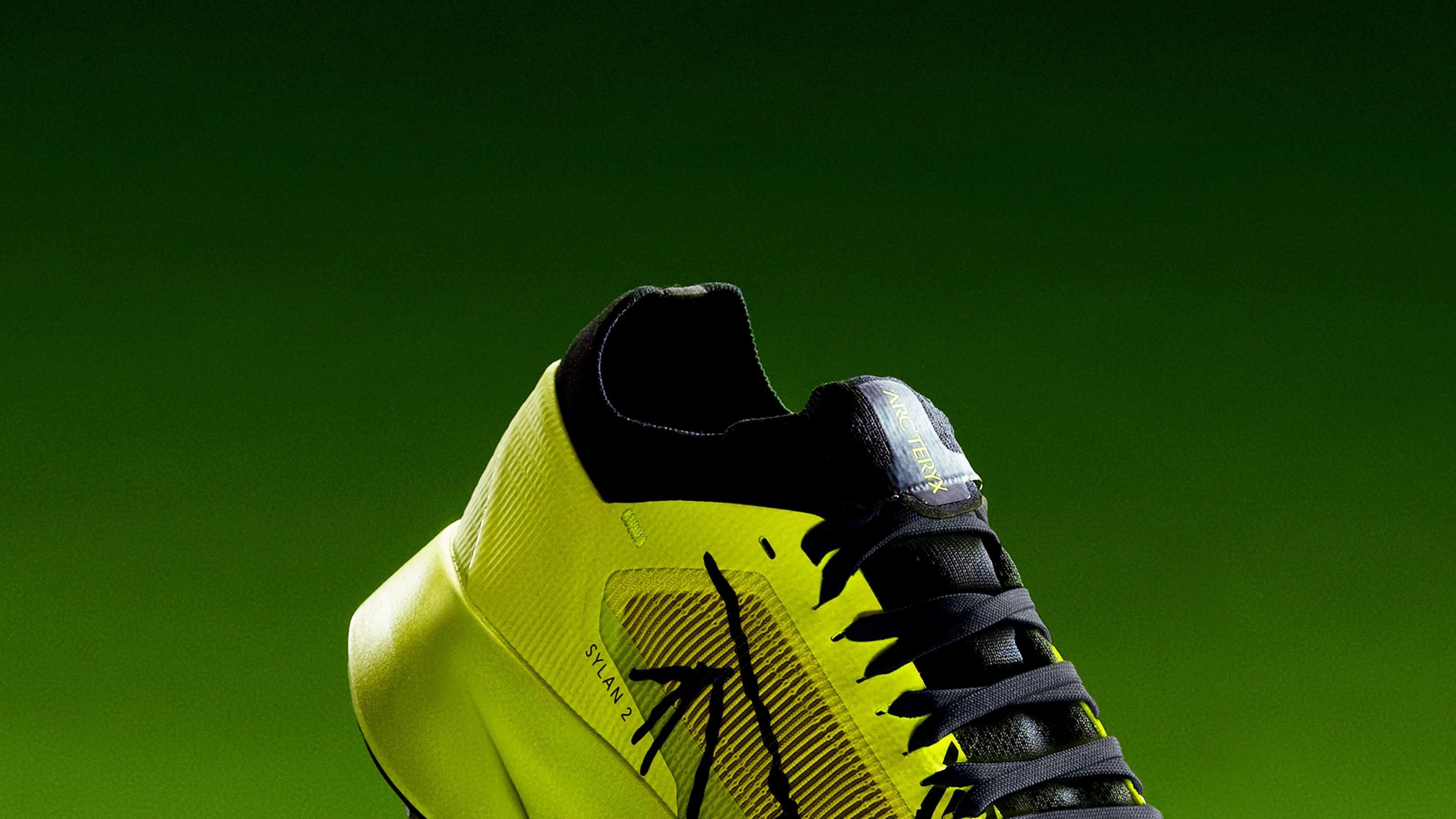 Yellow and black Arc'teryx Sylvan 2 running shoe with black laces on a green background.
