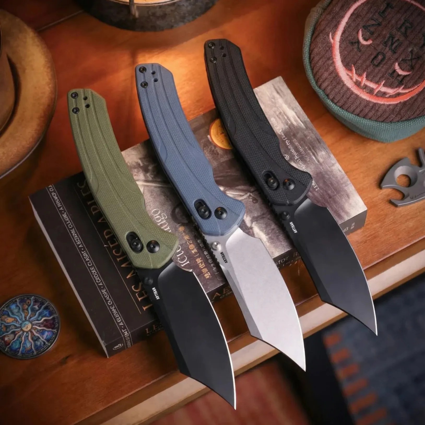Three folding knives with textured handles in green, blue, and black, placed on a wooden surface with books underneath.