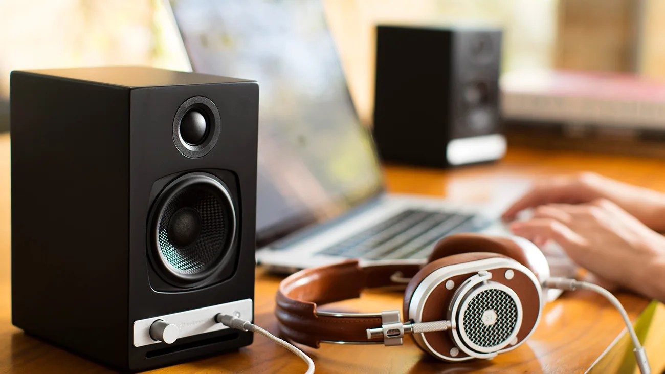 Black desktop speaker with silver volume knob and connected brown over-ear headphones on a wooden desk near a laptop.