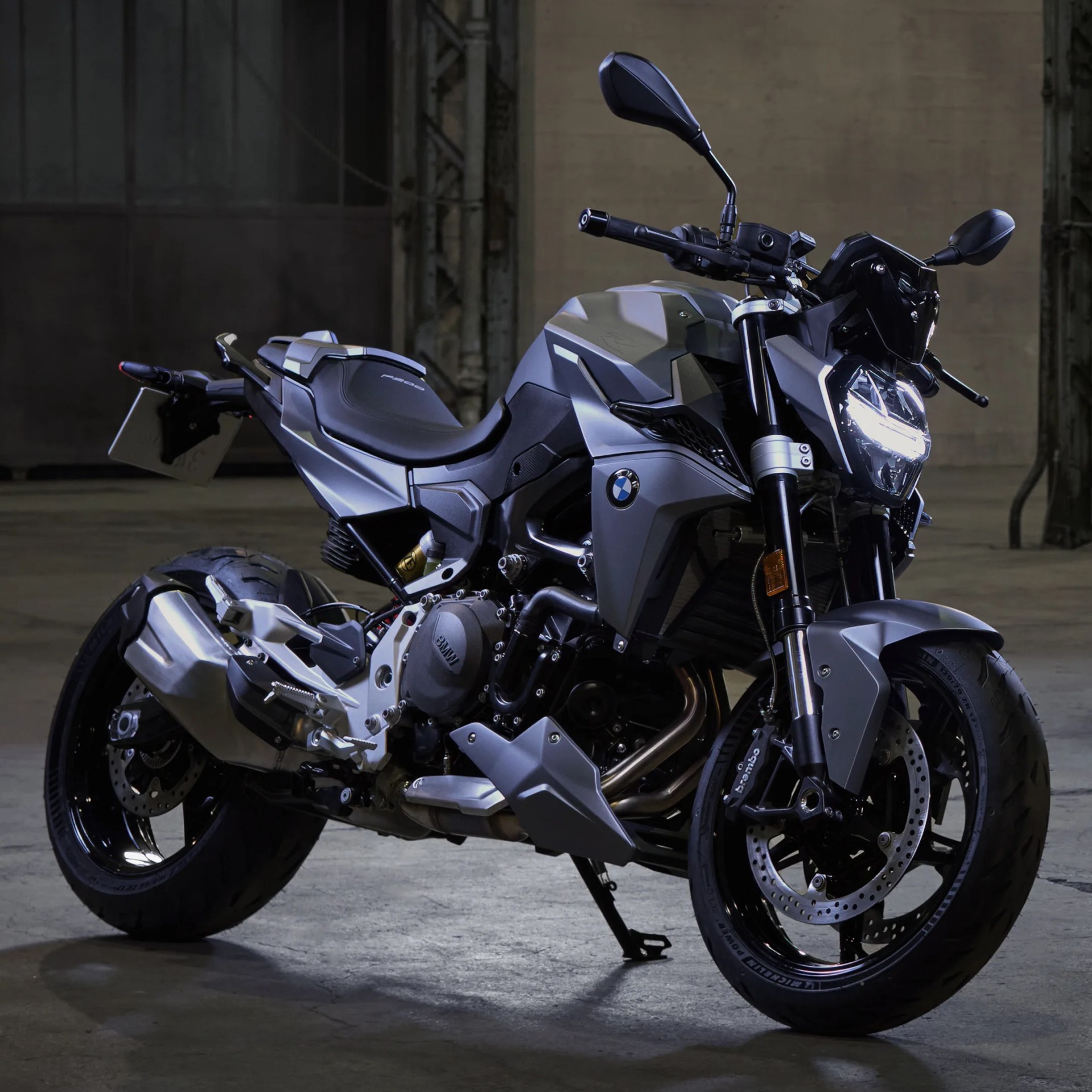 Matte gray BMW motorcycle with black accents and illuminated headlight in a dim industrial setting.