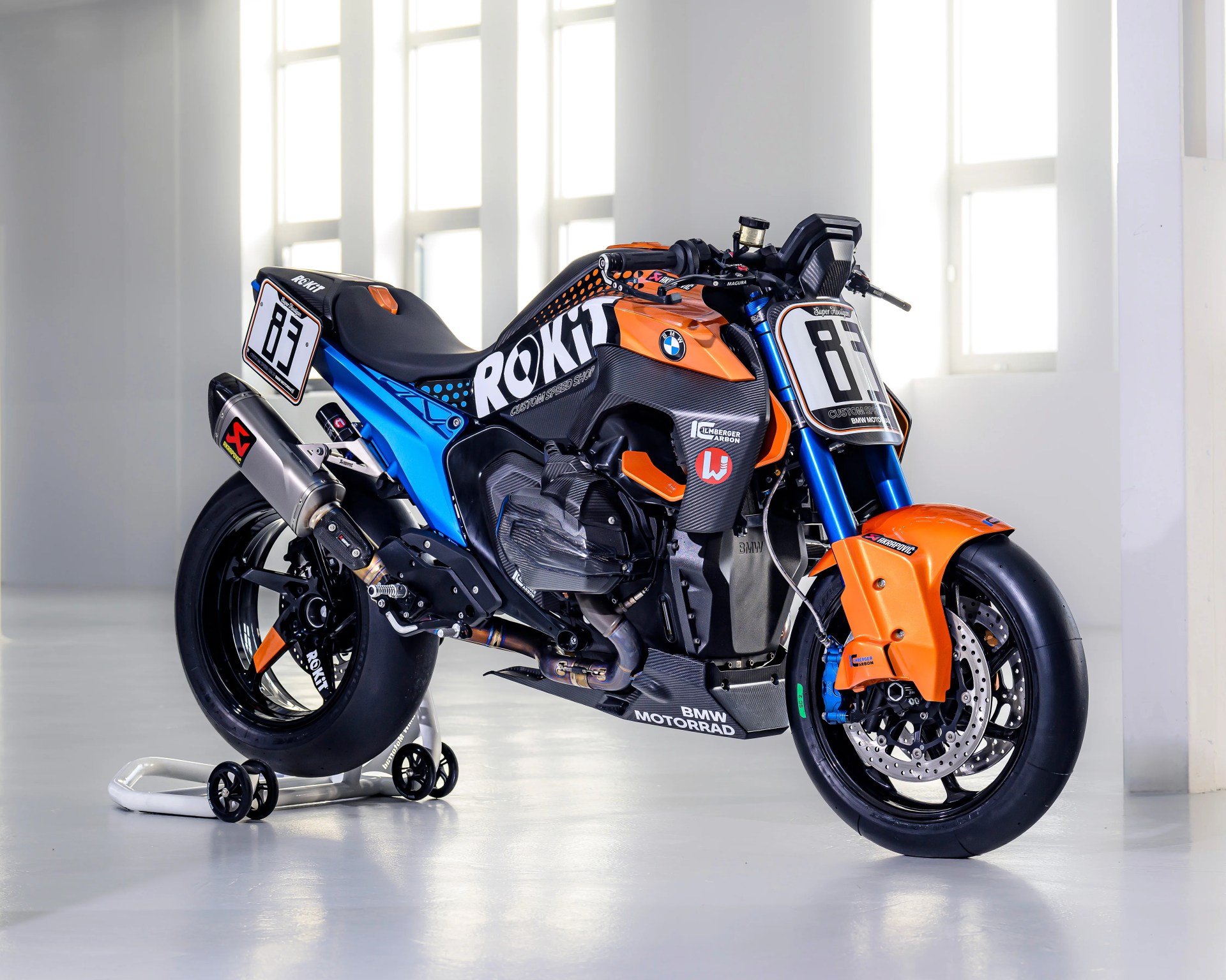 Orange and blue BMW Motorrad racing motorcycle with ROKiT branding on a white stand in a bright room.