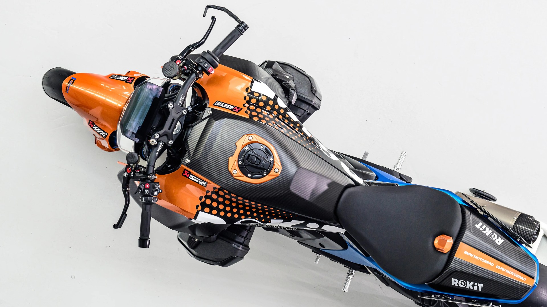Top-down view of a black, orange, and blue sport motorcycle with Akrapovic and ROKiT branding on a white background.