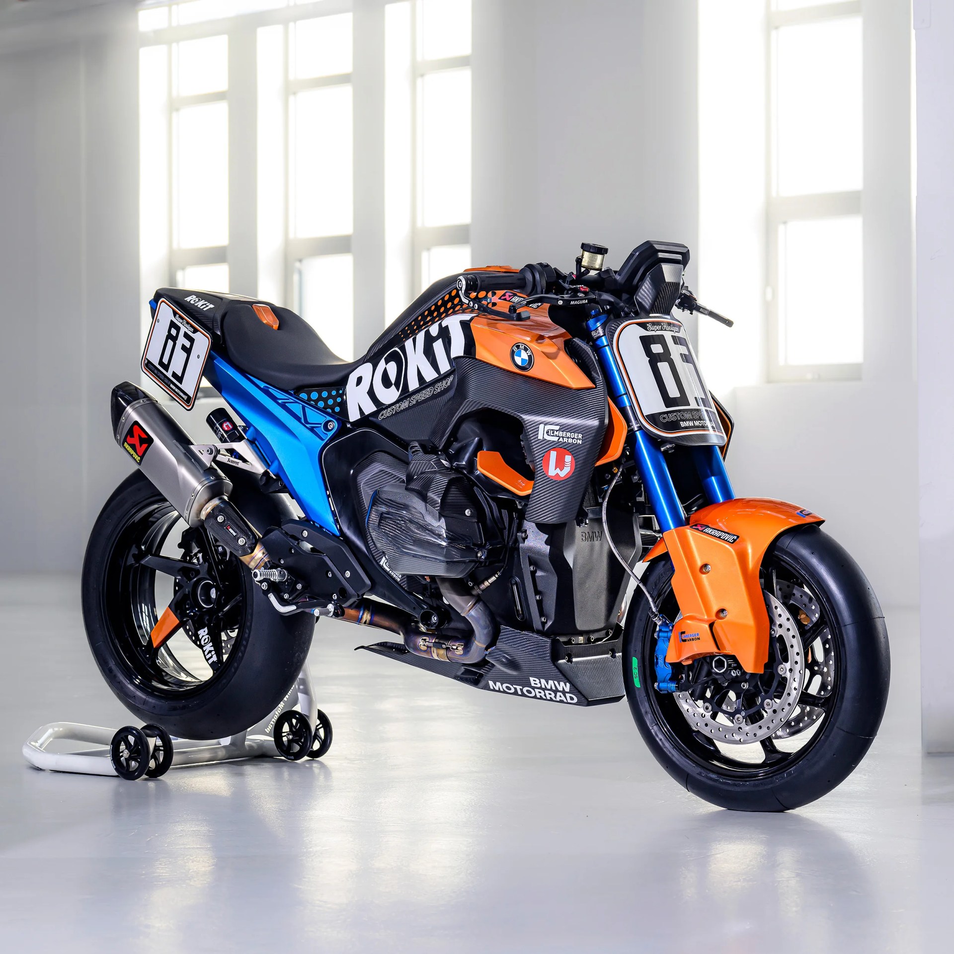 Rokit-branded BMW Motorrad racing motorcycle with orange, blue, and black carbon fiber body on a white stand.