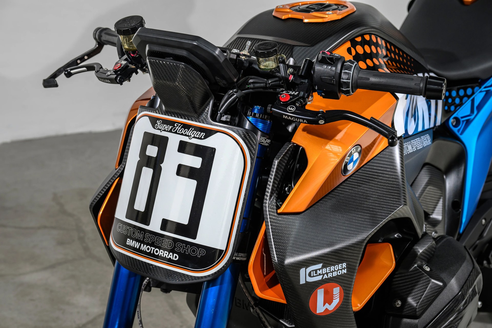 Close-up of a BMW motorcycle front with orange and blue carbon fiber bodywork, number plate "83," and Magura brake lever.