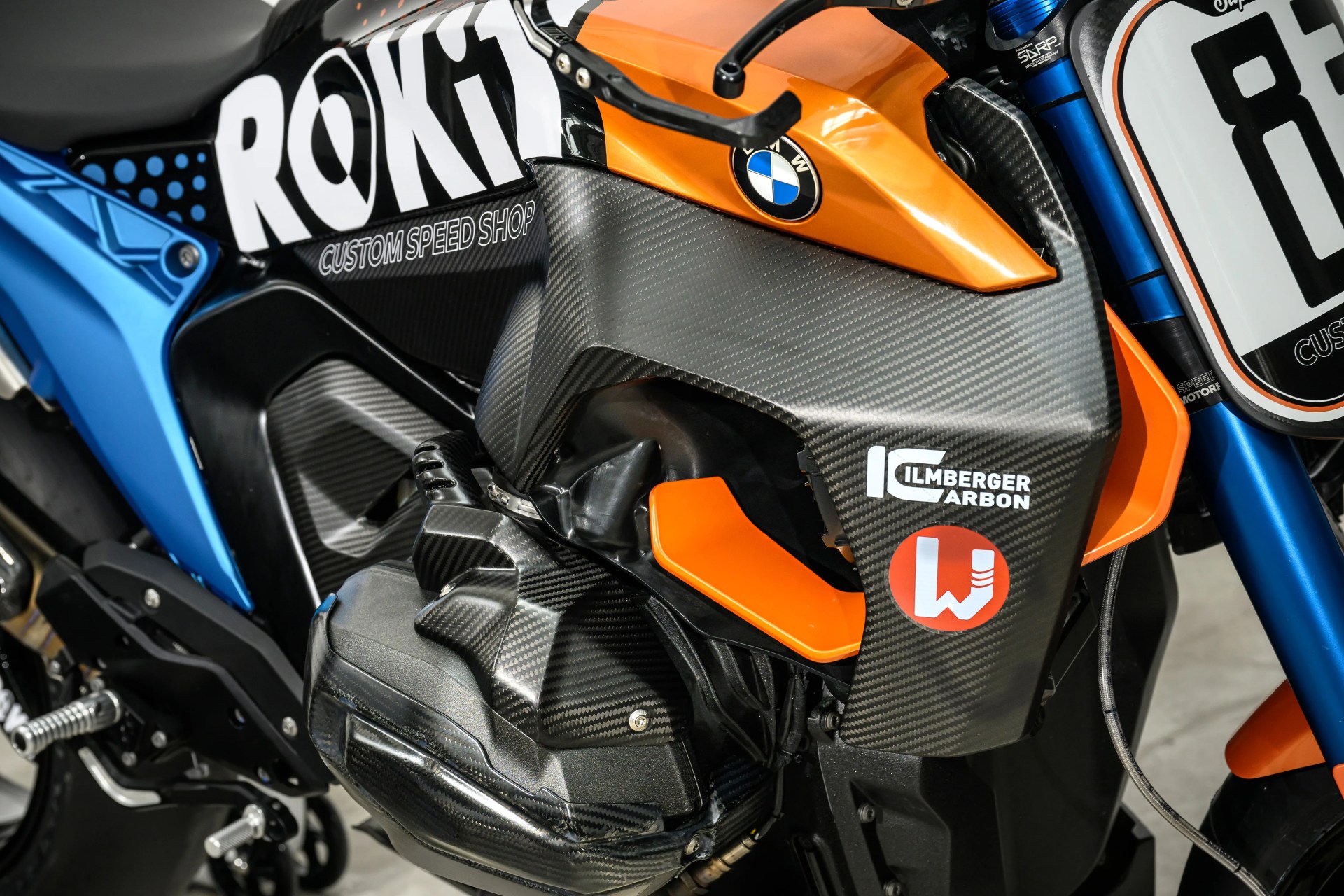 Close-up of a BMW motorcycle engine with carbon fiber and orange body panels, featuring ROKiT and Ilmberger Carbon logos.
