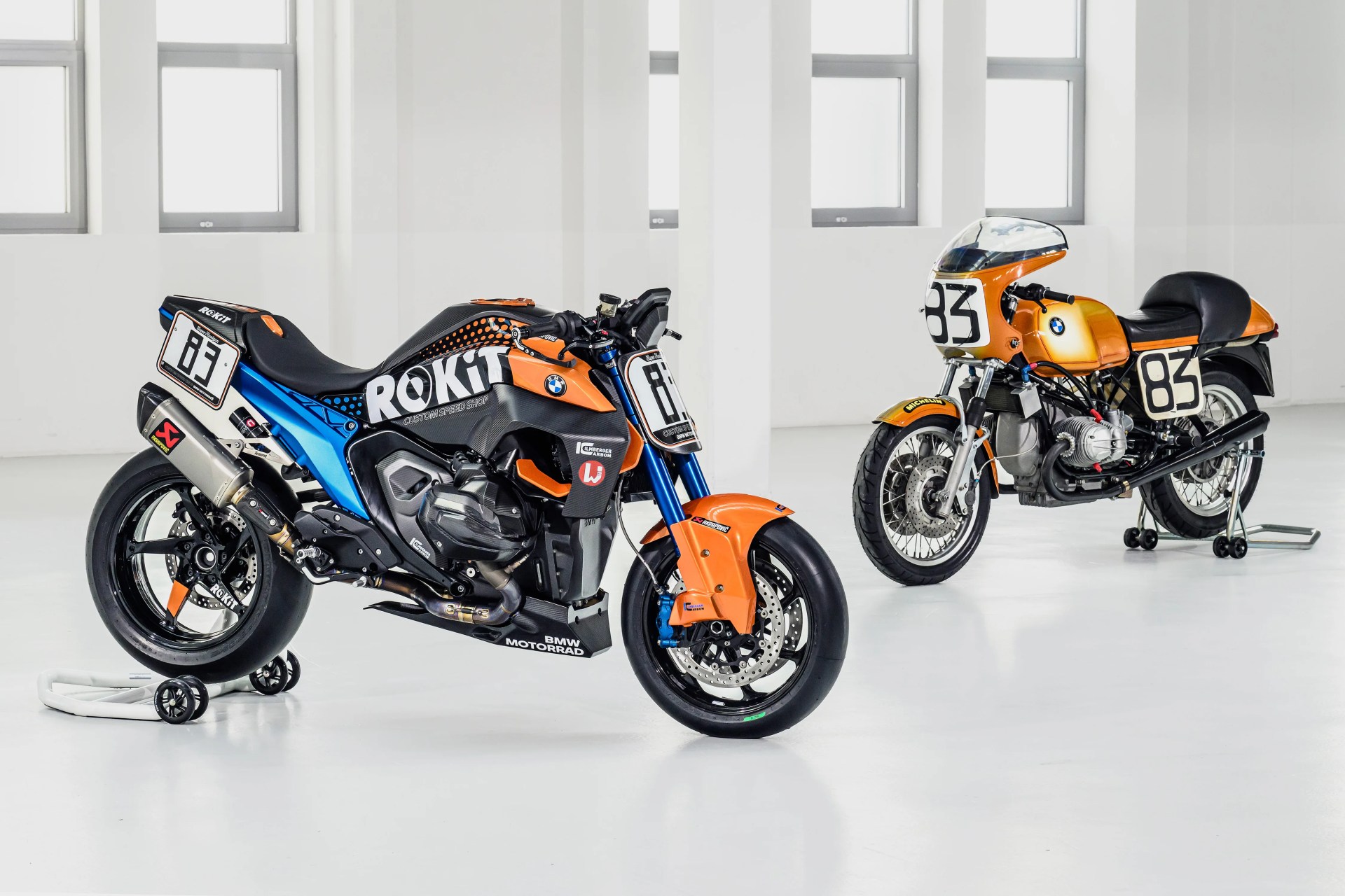 Two BMW motorcycles with orange and black racing designs, one modern and one vintage, displayed indoors on white stands.
