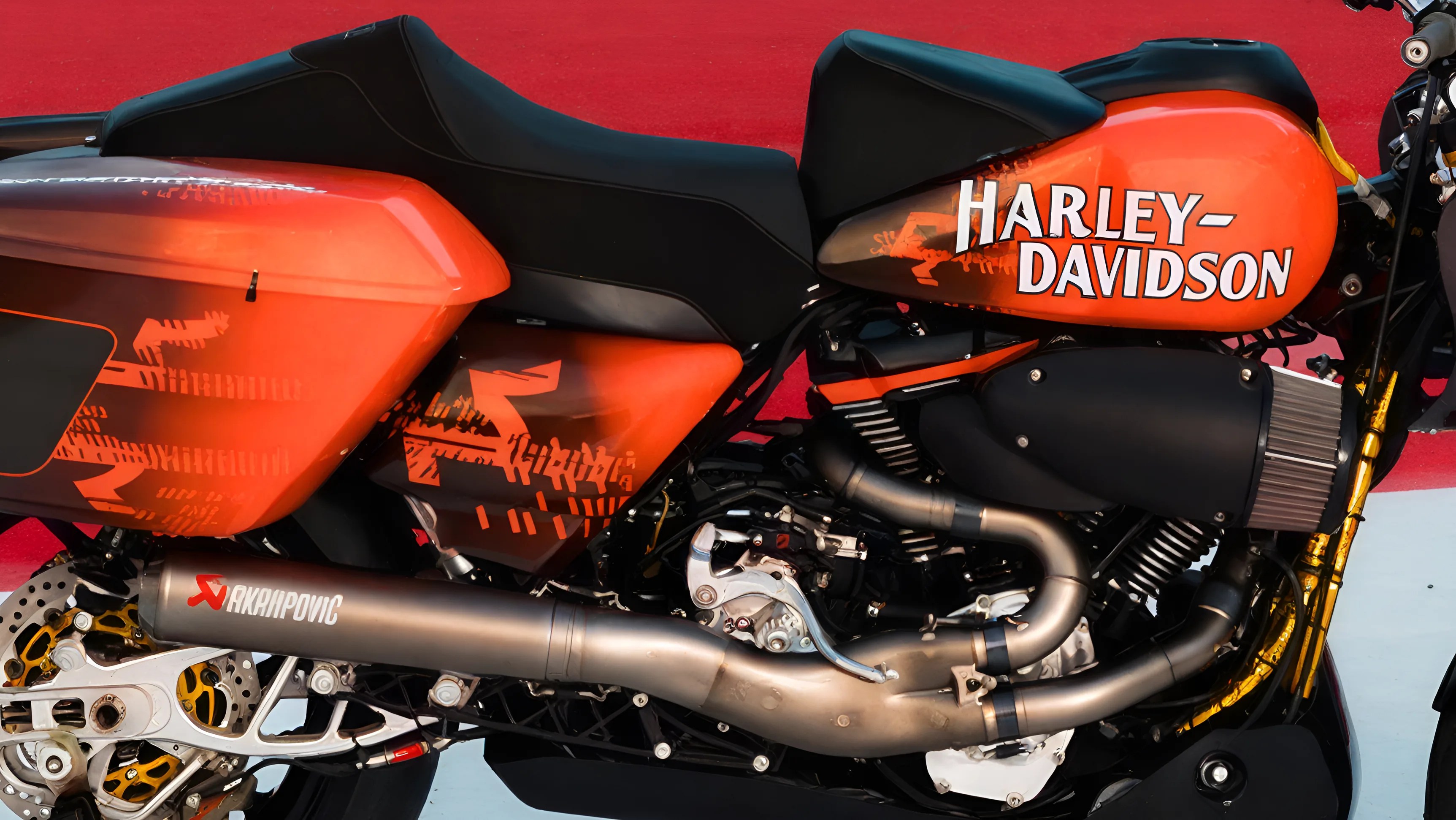 Orange and black Harley-Davidson motorcycle with Akrapovic exhaust and black seat on red and white surface.