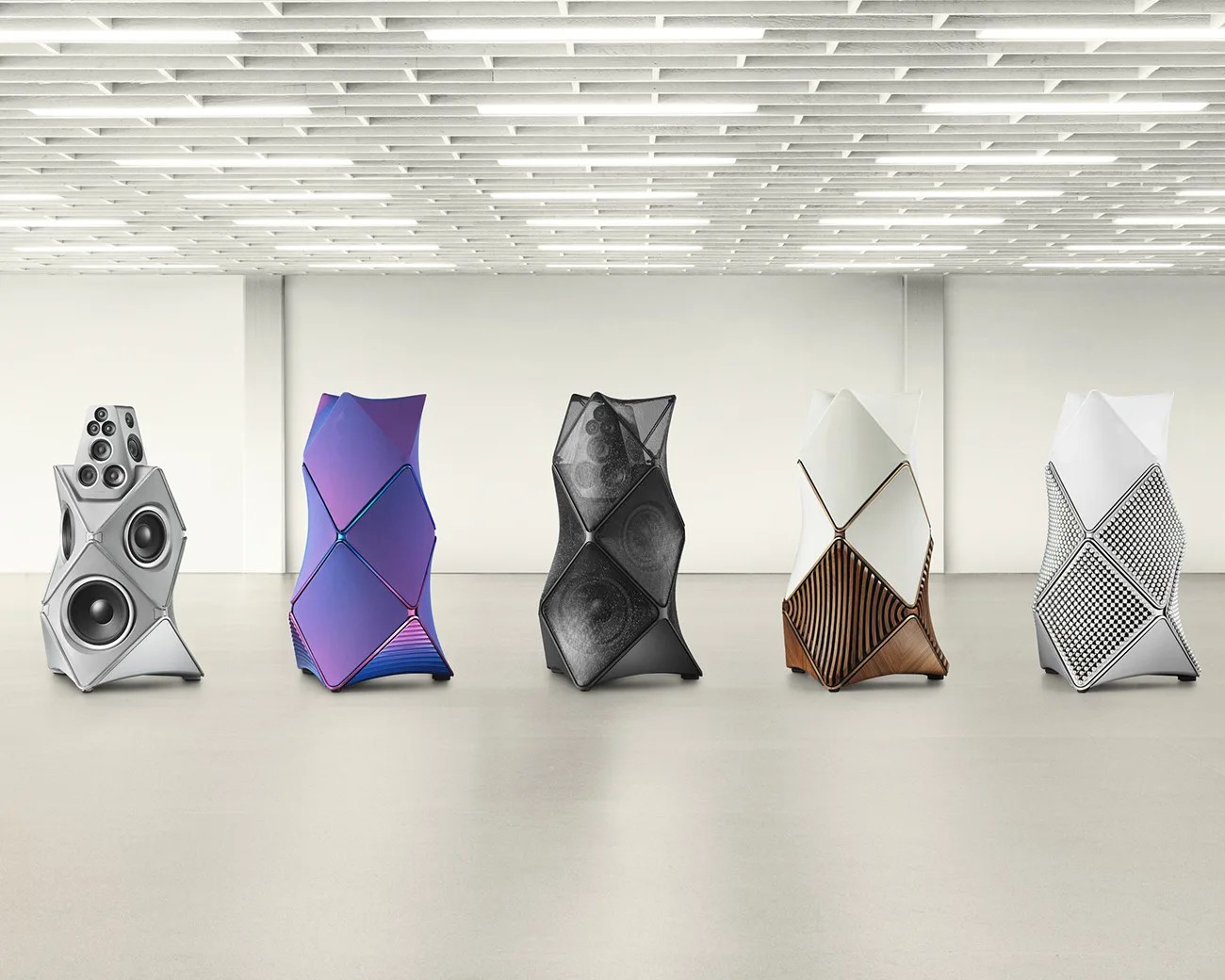 Five uniquely designed modern speakers with geometric, faceted exteriors in silver, purple, black, white with wood, and white with patterned fabric, displayed in a minimalist room.