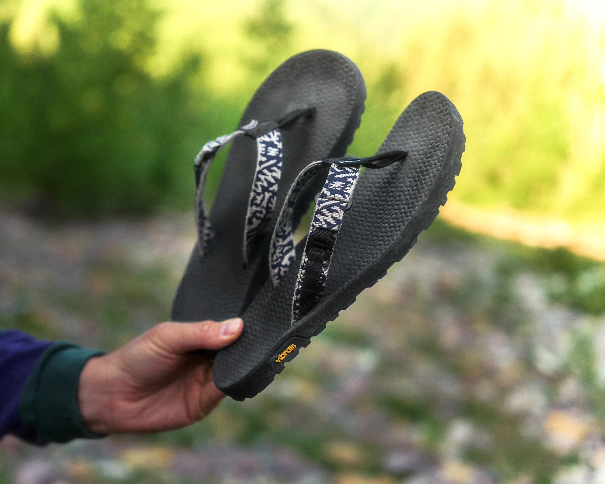 Black textured flip-flops with patterned fabric straps held in a hand outdoors.