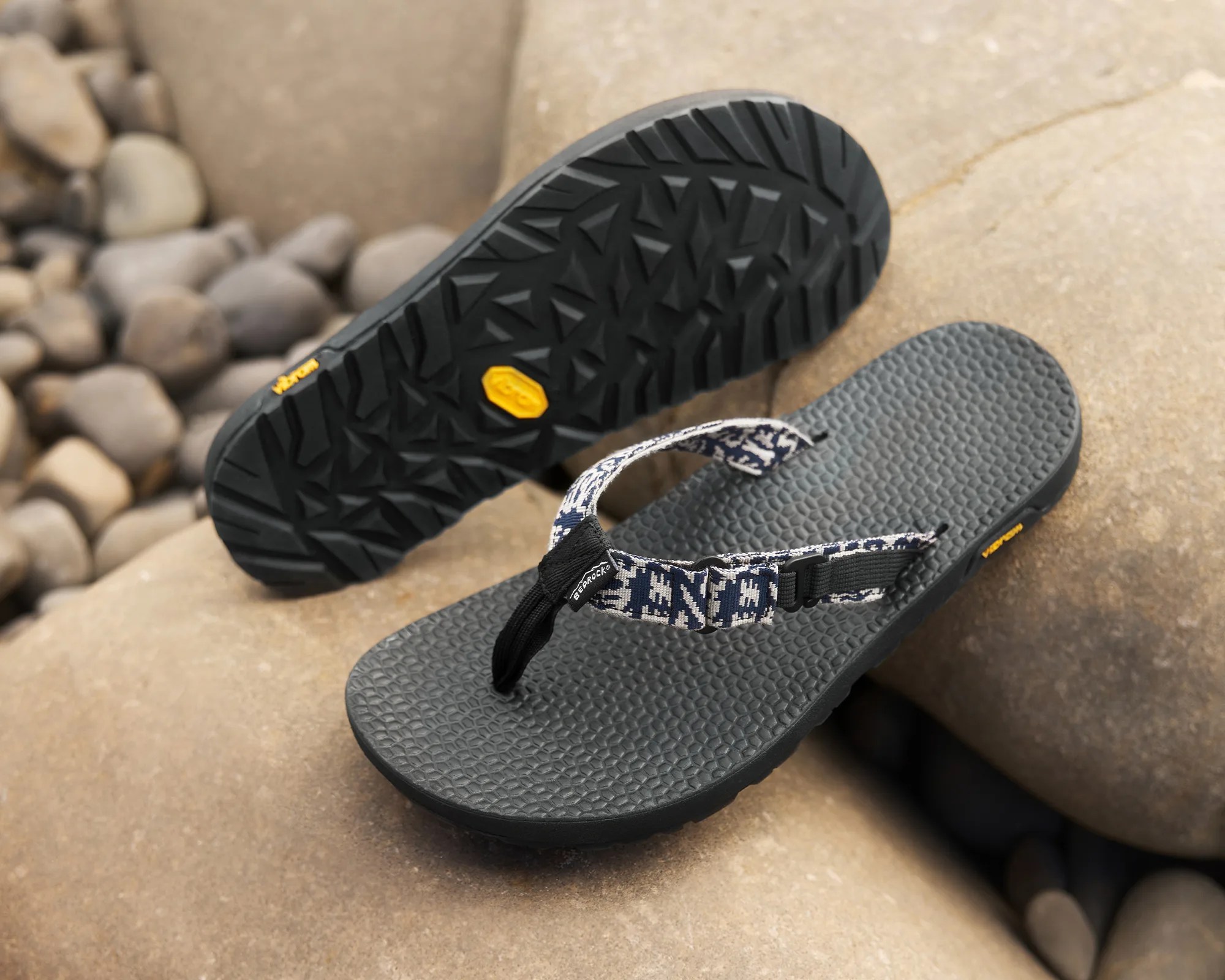 Pair of black textured sole flip-flops with blue and white patterned fabric straps on rocks.