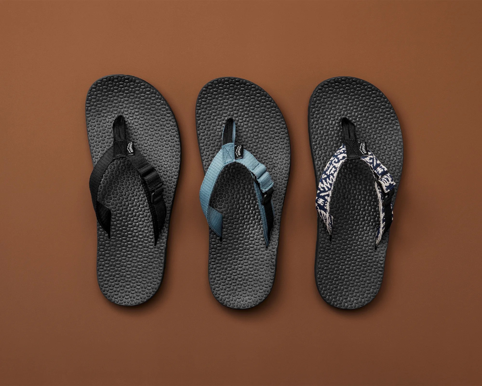 Three black textured flip-flops with different colored fabric straps&mdash;black, blue, and navy with white text&mdash;on a brown background.