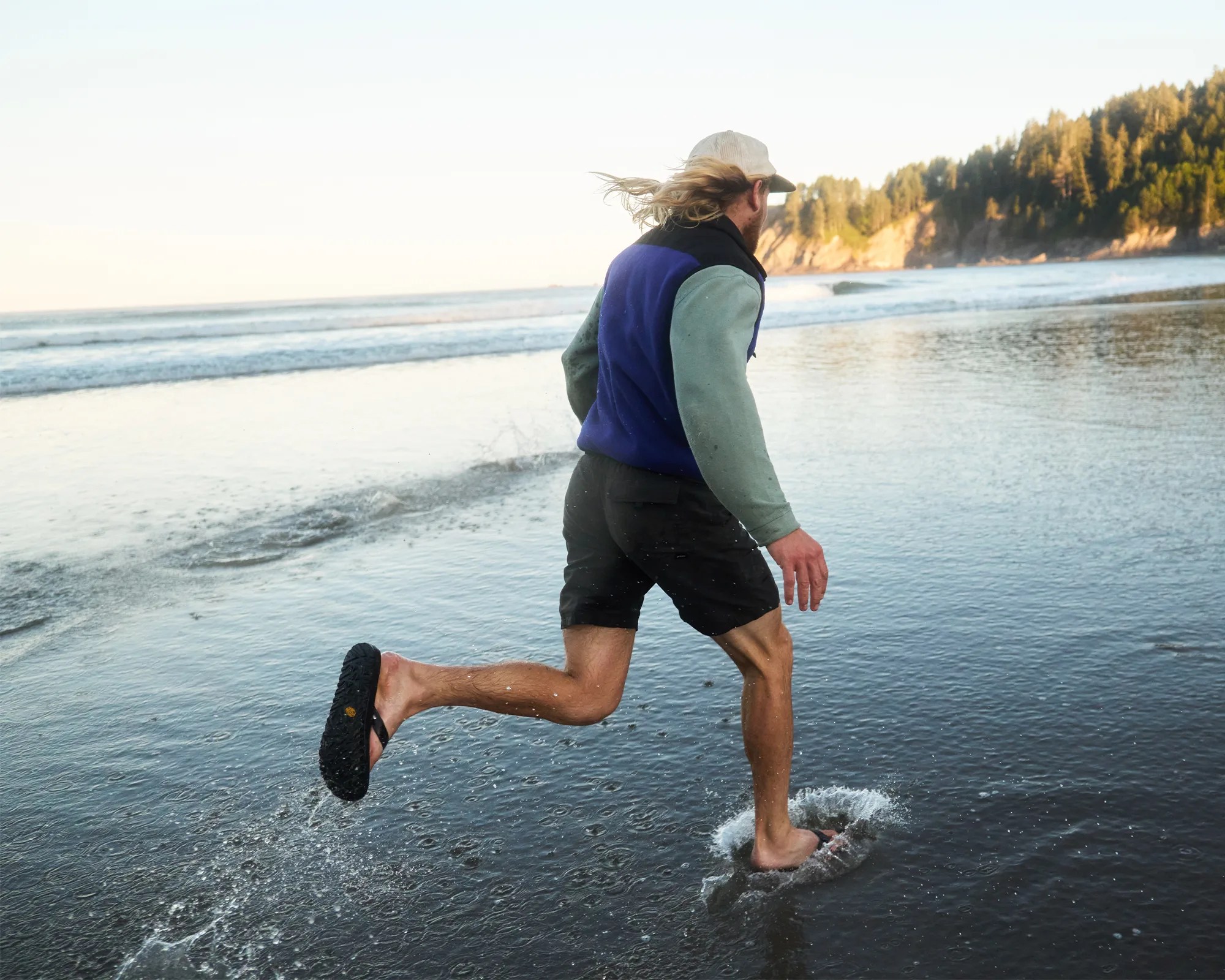 Person wearing a beige cap, blue and green jacket, and black shorts running barefoot on a wet beach.