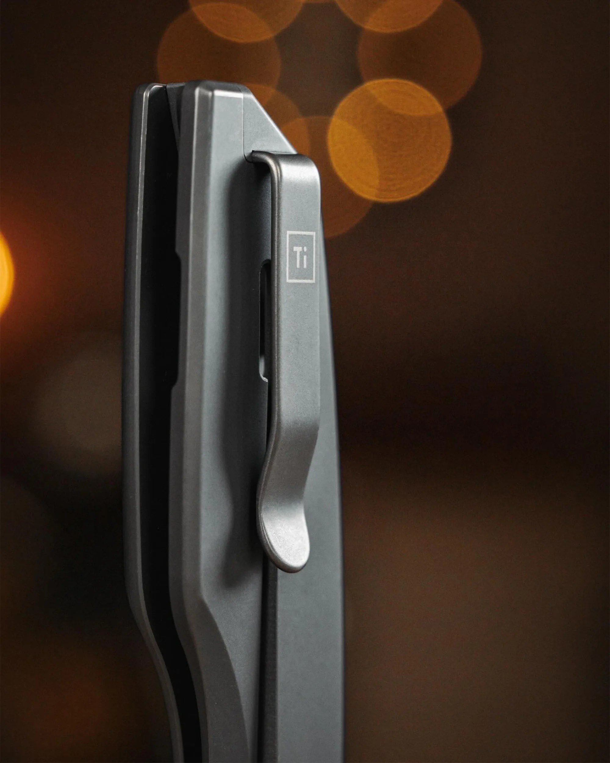 Close-up of a gray titanium pocket clip attached to a folded knife handle with a blurred warm light background.