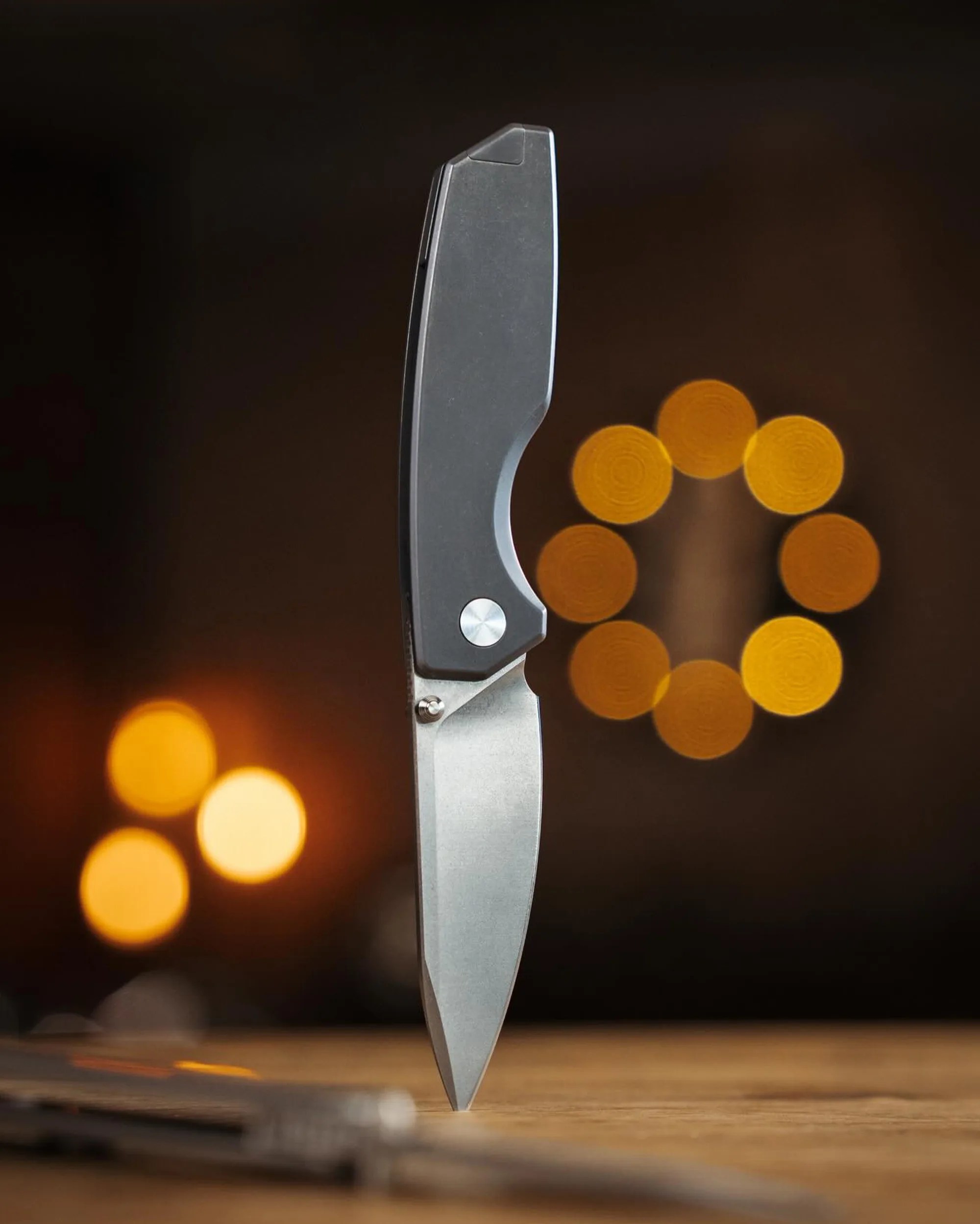 Folding knife with a matte black handle and silver blade standing upright on a wooden surface with blurred warm lights in the background.