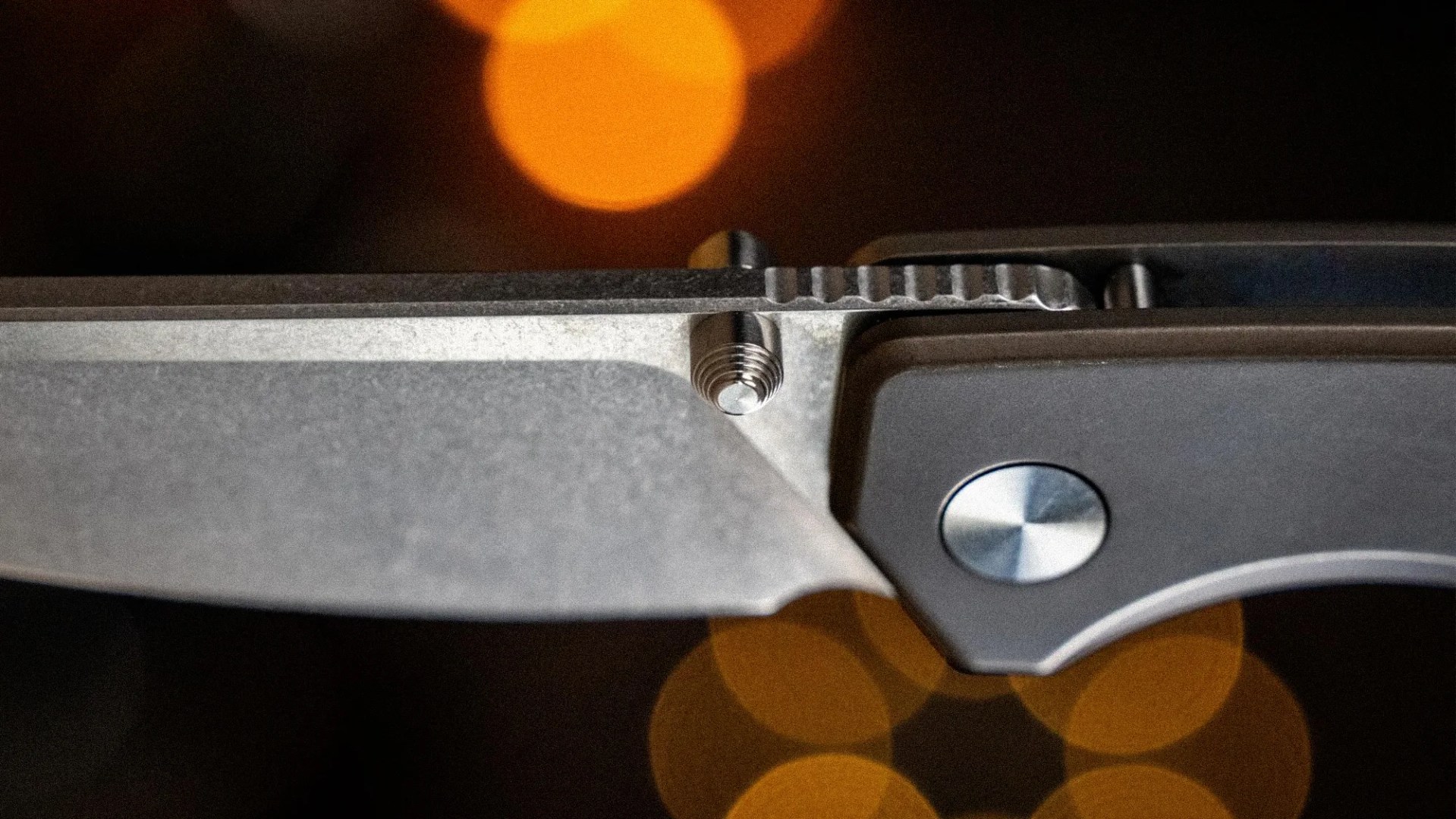 Close-up of a partially open folding knife with a stonewashed blade and a gray handle against a dark background with orange bokeh lights.