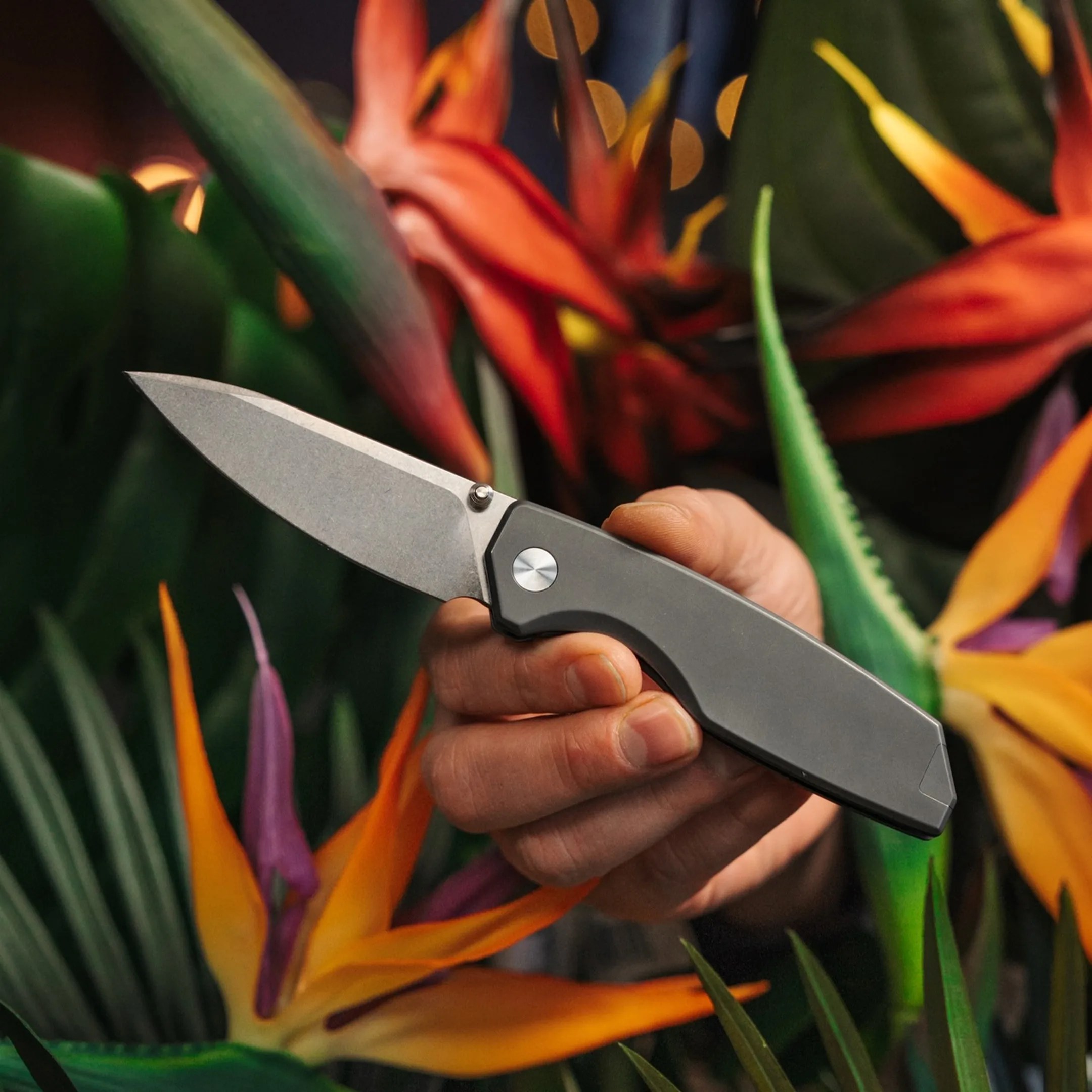 Hand holding a gray folding knife with a stonewashed blade among colorful tropical flowers.