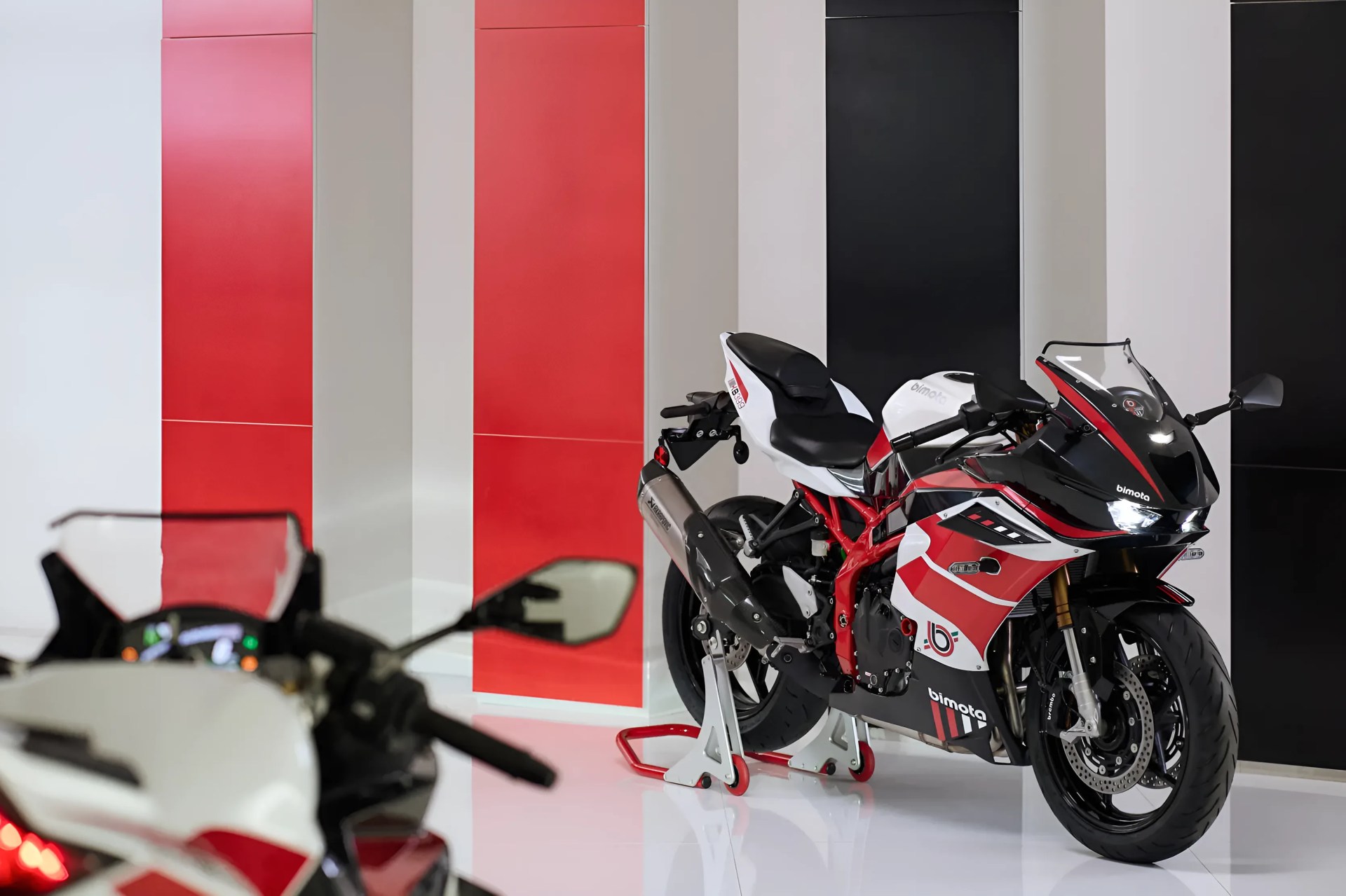 Red, white, and black Bimota sport motorcycle on a white stand in a showroom with red and black wall panels.