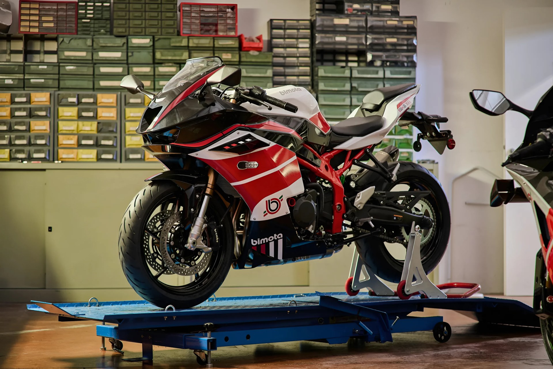 Red, white, and black Bimota sport motorcycle on a blue maintenance lift in a workshop with organized drawers in the background.