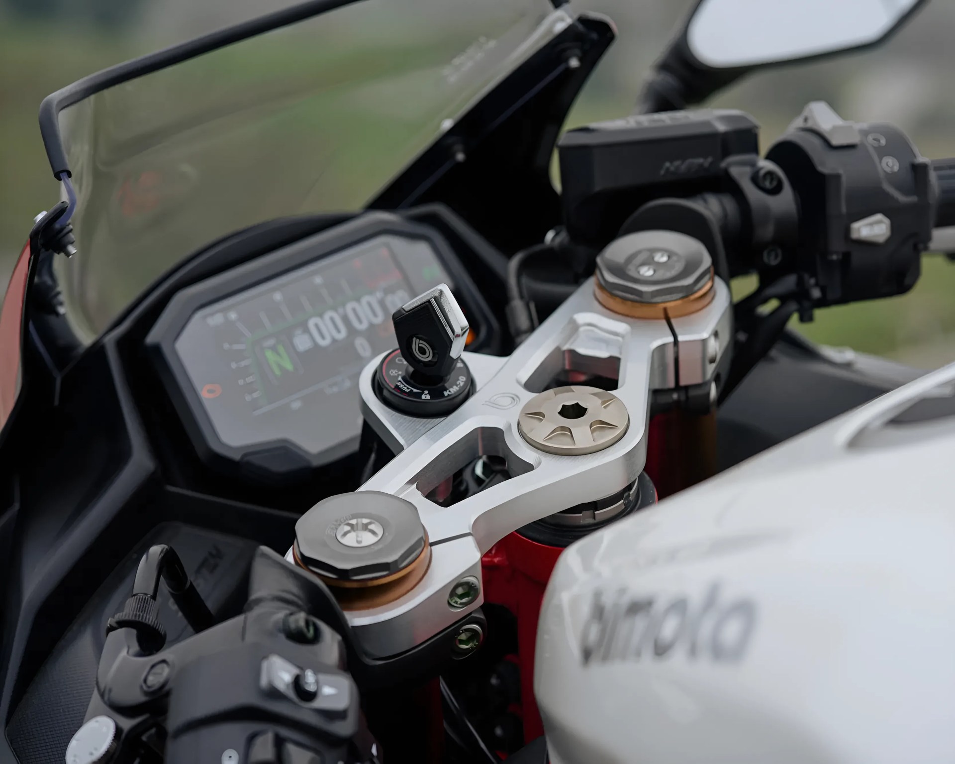 Close-up of a motorcycle handlebar with a digital dashboard, silver triple clamp, and control switches.