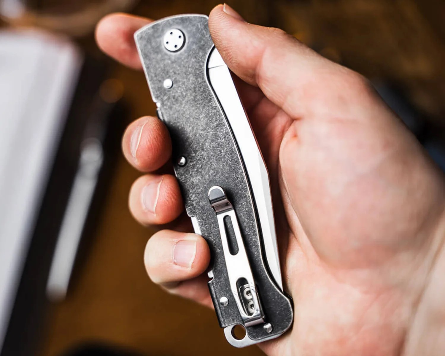 Hand holding a closed folding knife with a stonewashed metal handle and a silver pocket clip.