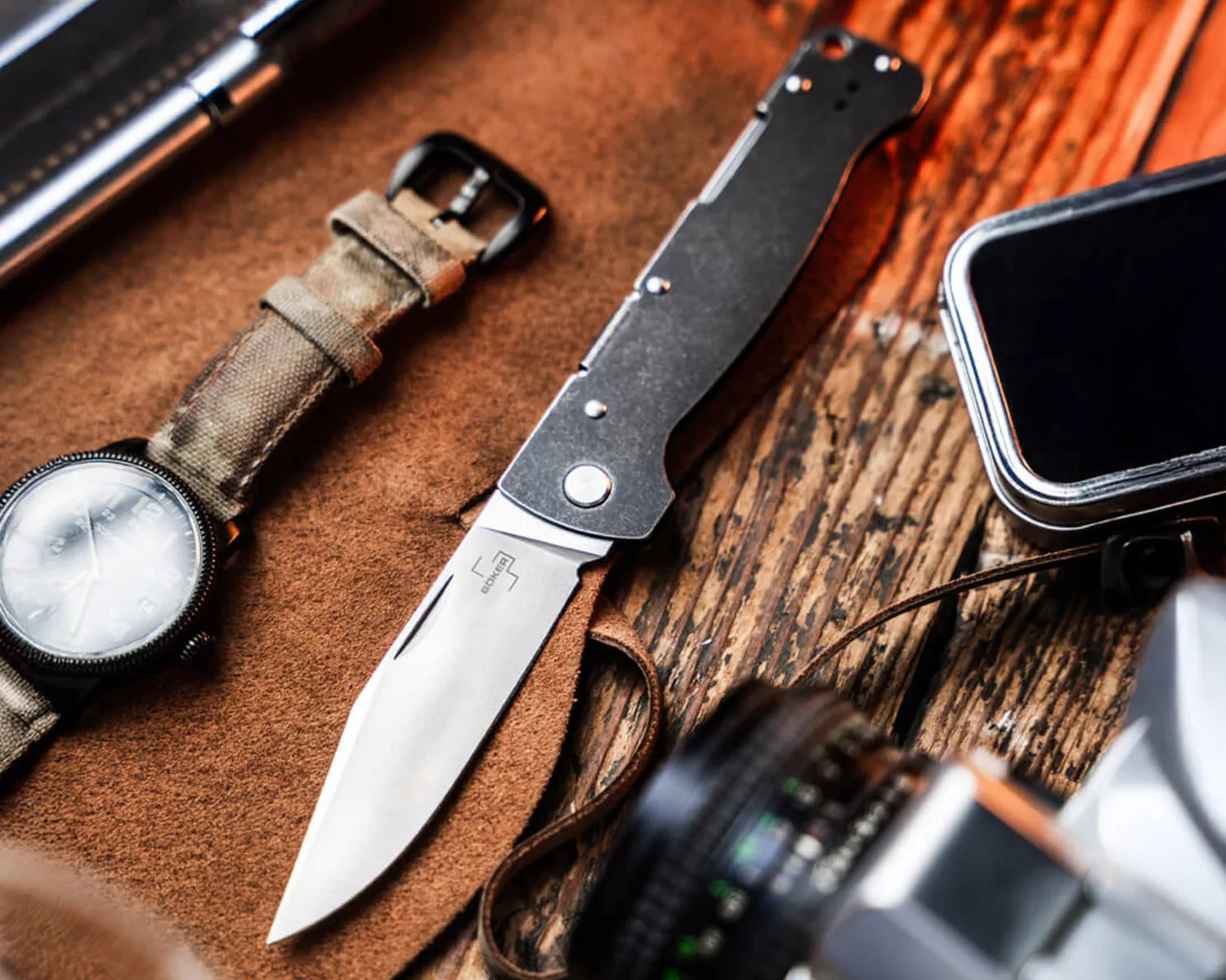 Folding knife with black handle and silver blade on a brown leather surface next to a watch and camera.